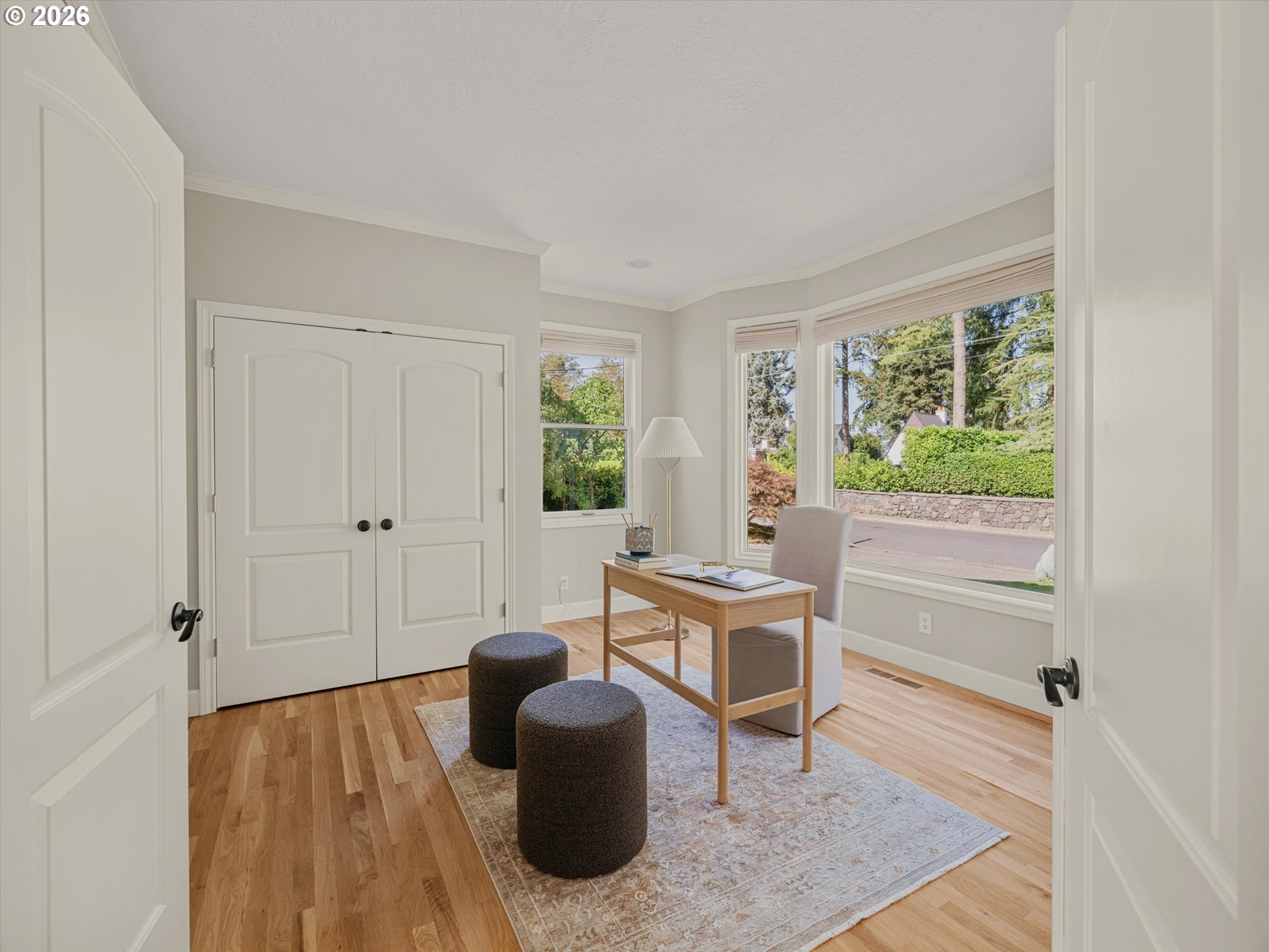 Bright home office with hardwood floors, white closet doors, and large windows overlooking greenery.