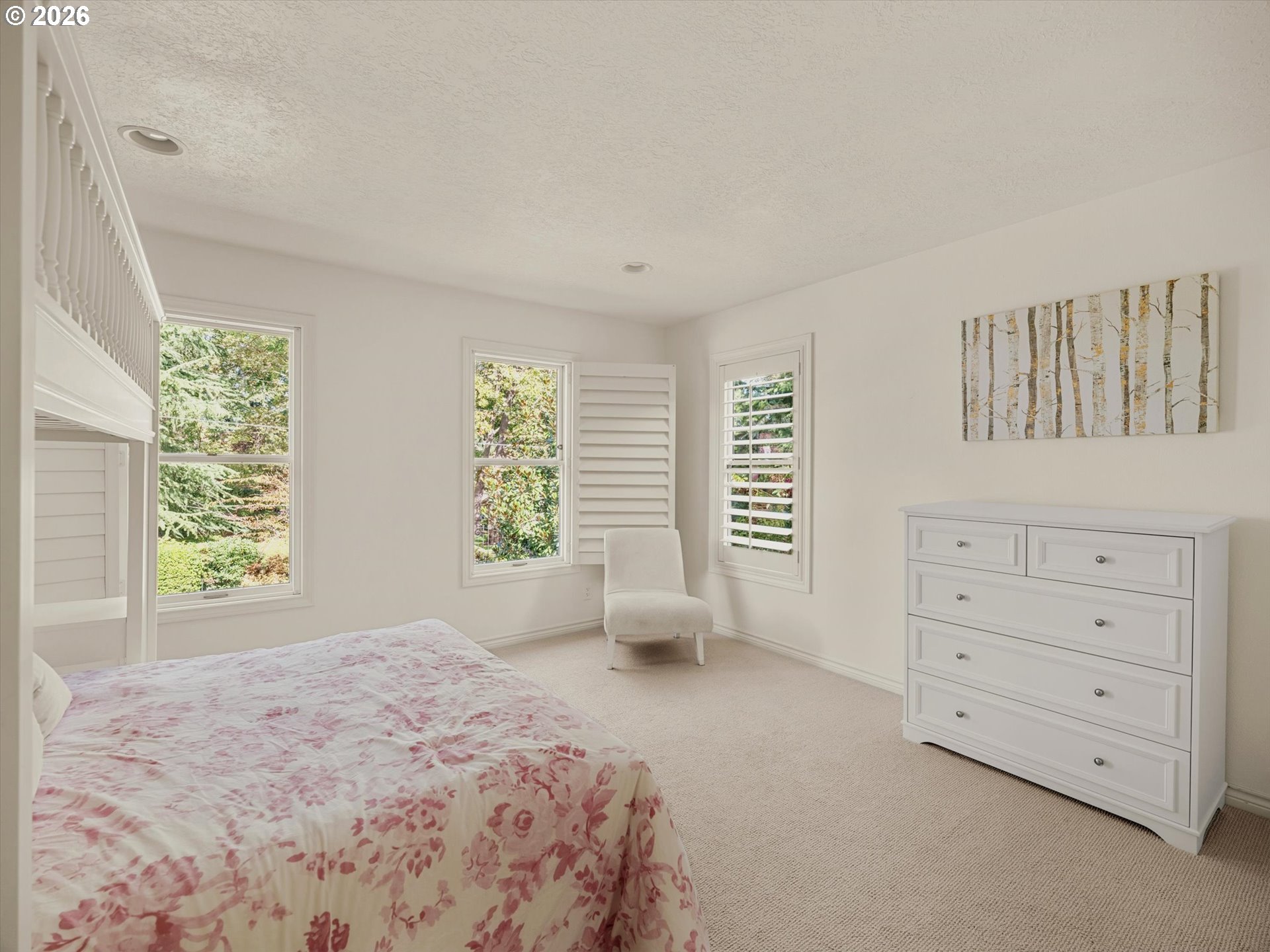 Bright bedroom with floral bedspread, white dresser, and shuttered windows overlooking greenery.