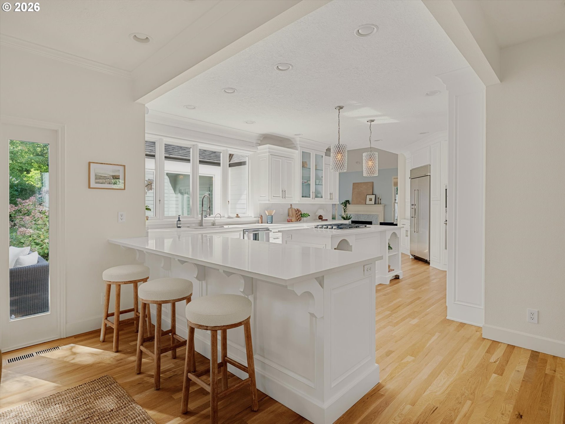 Bright white kitchen with a large island, three stools, pendant lights, and light hardwood floors.