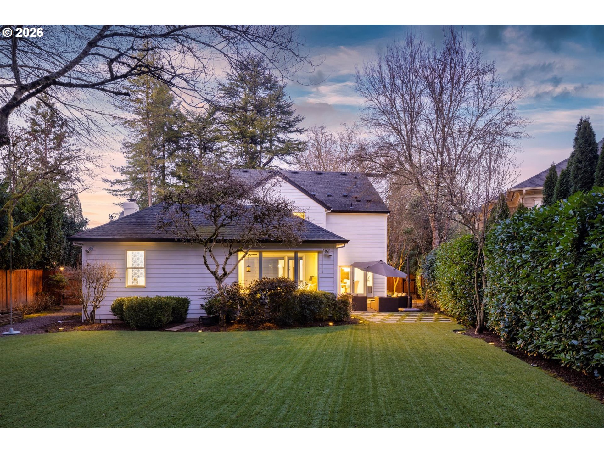 House rear exterior with illuminated windows, a manicured lawn, and a patio umbrella at dusk.