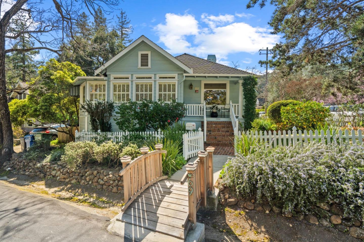Pale green cottage with a covered porch, white picket fence, and wooden entry bridge.