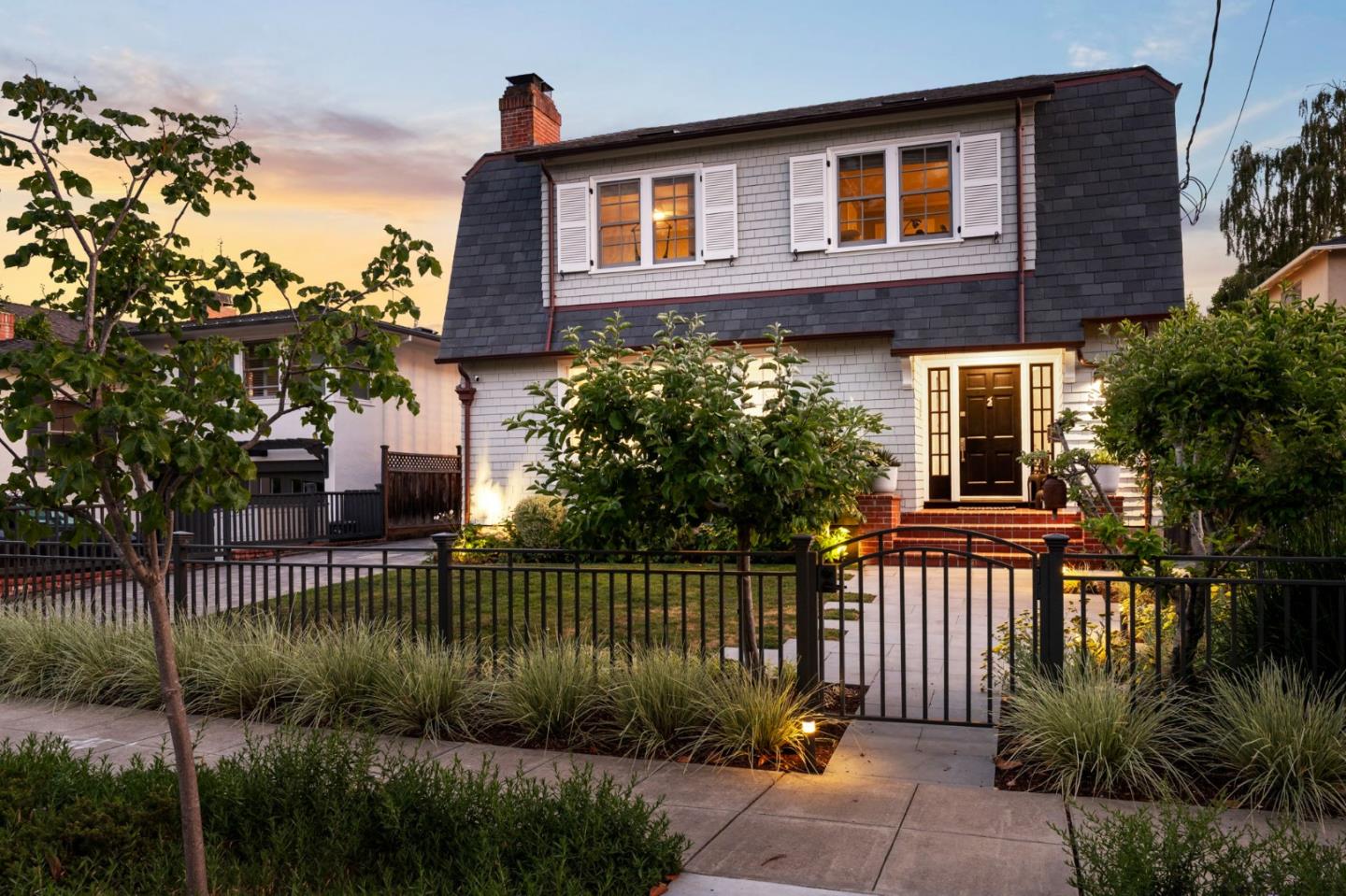 Two-story shingle-sided home with white shutters, a black metal front gate, and warmly lit windows at dusk.