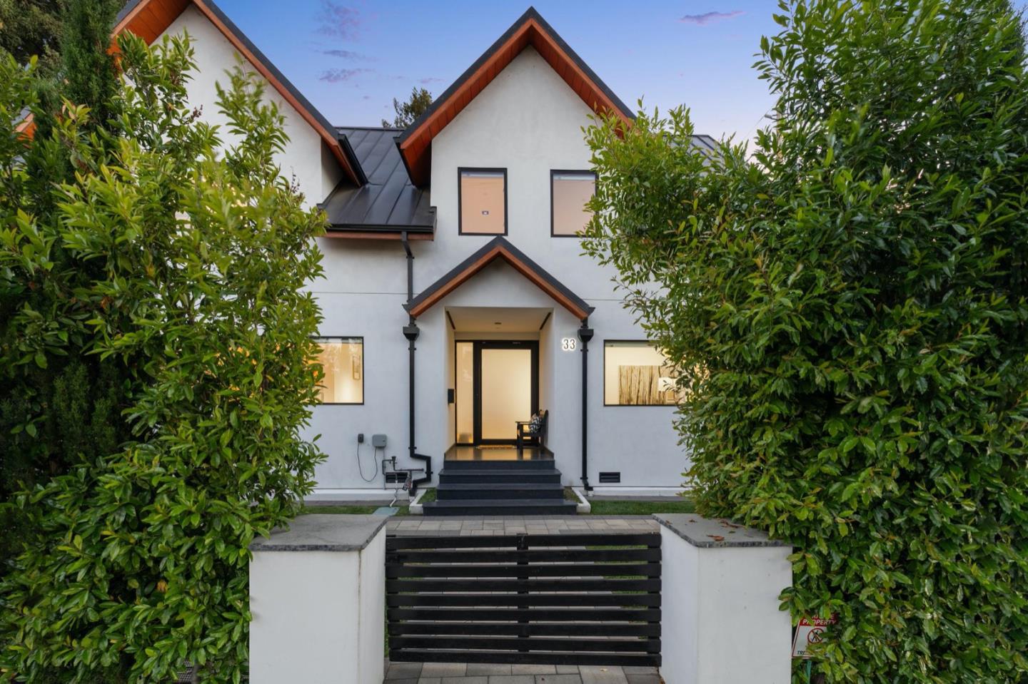 Modern white two-story house with wood-trimmed gables, framed by tall green hedges.