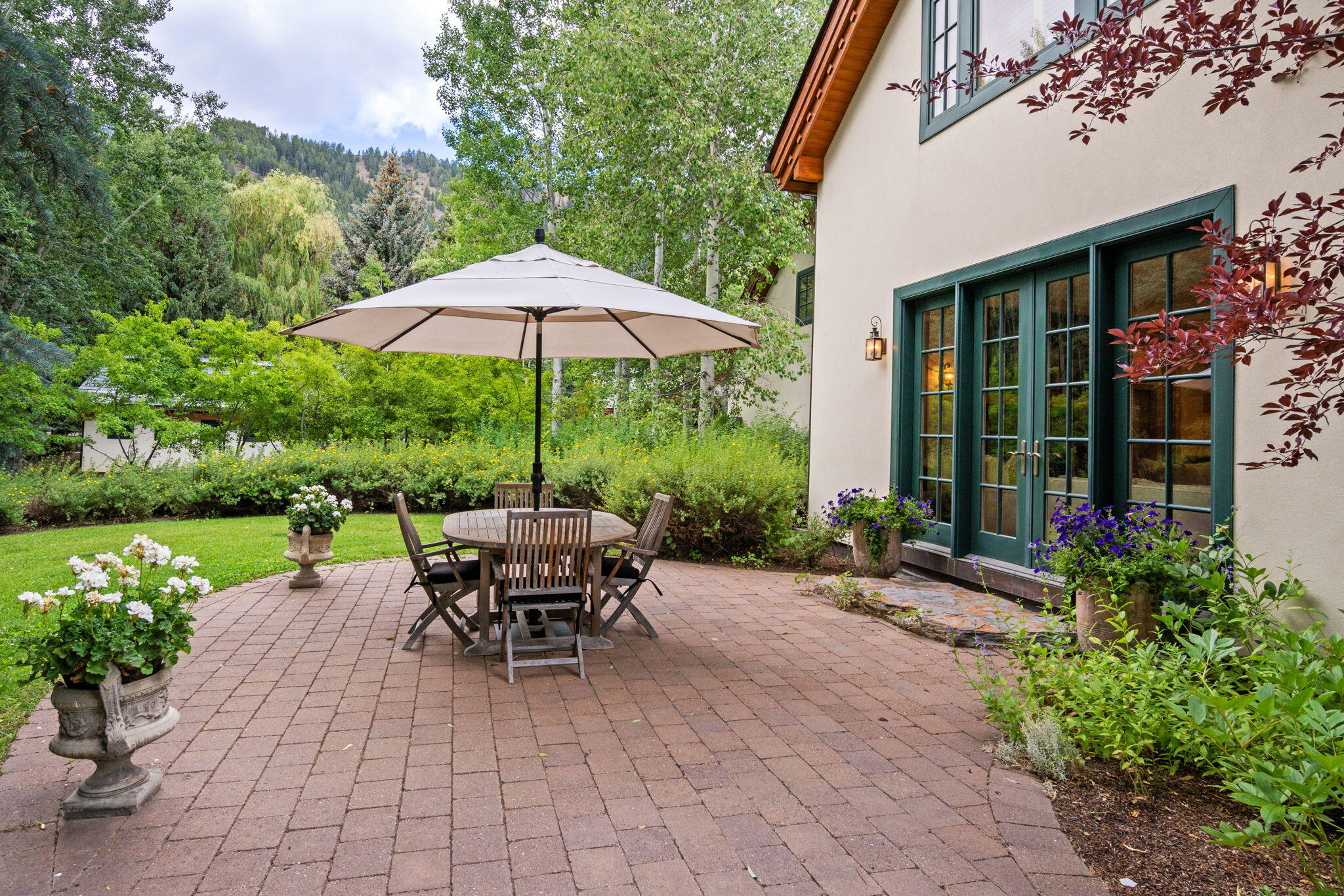 Brick patio with umbrella-shaded dining set beside French doors, surrounded by flowers and mature trees.