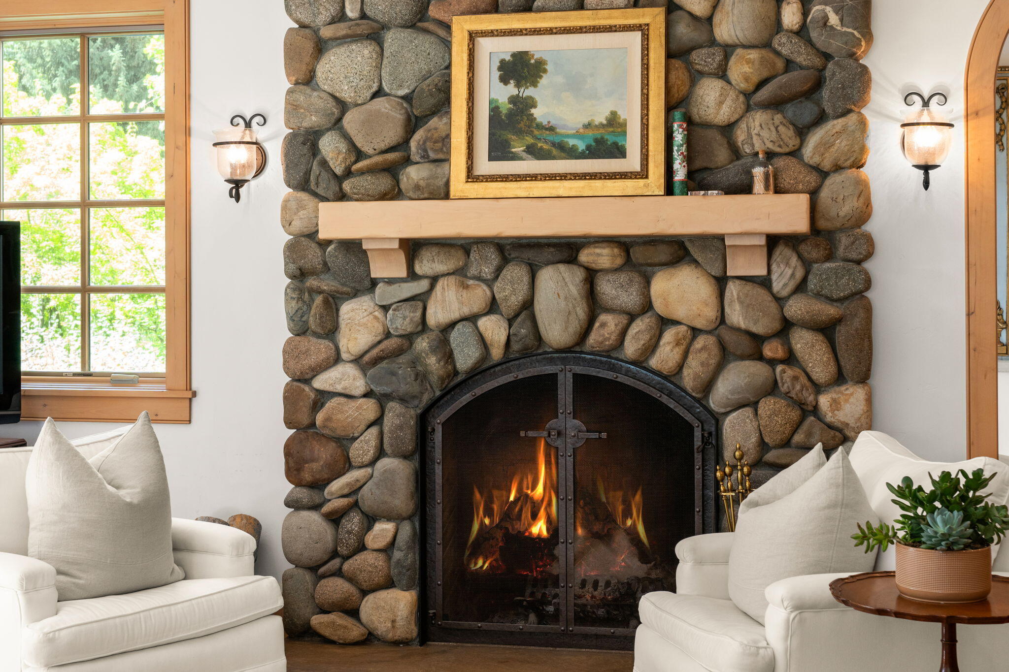 Living room with a river-stone fireplace, burning fire, and white upholstered chairs.