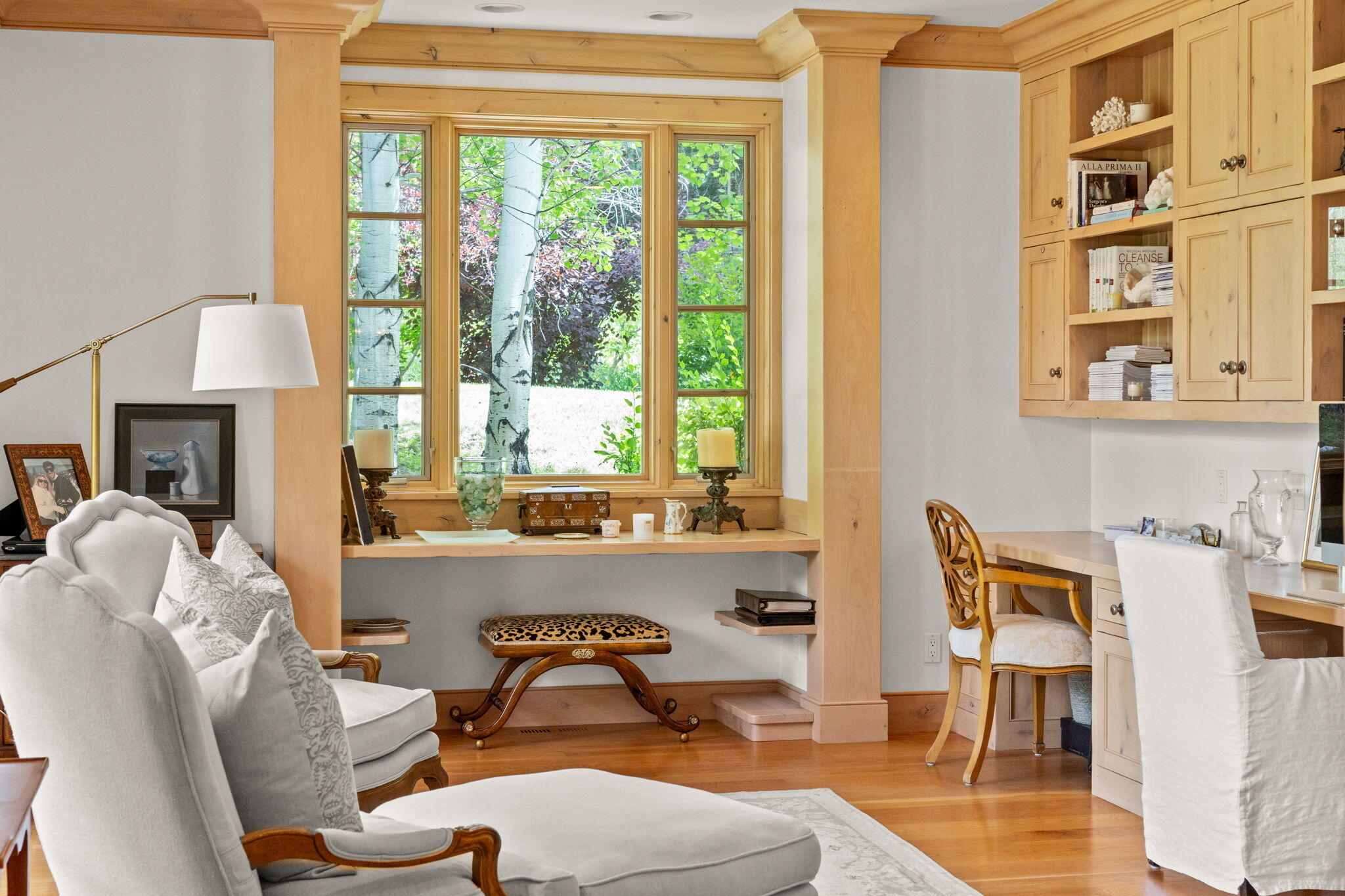 Furnished home office with built-in wood desk and cabinetry beside a large window overlooking leafy trees.