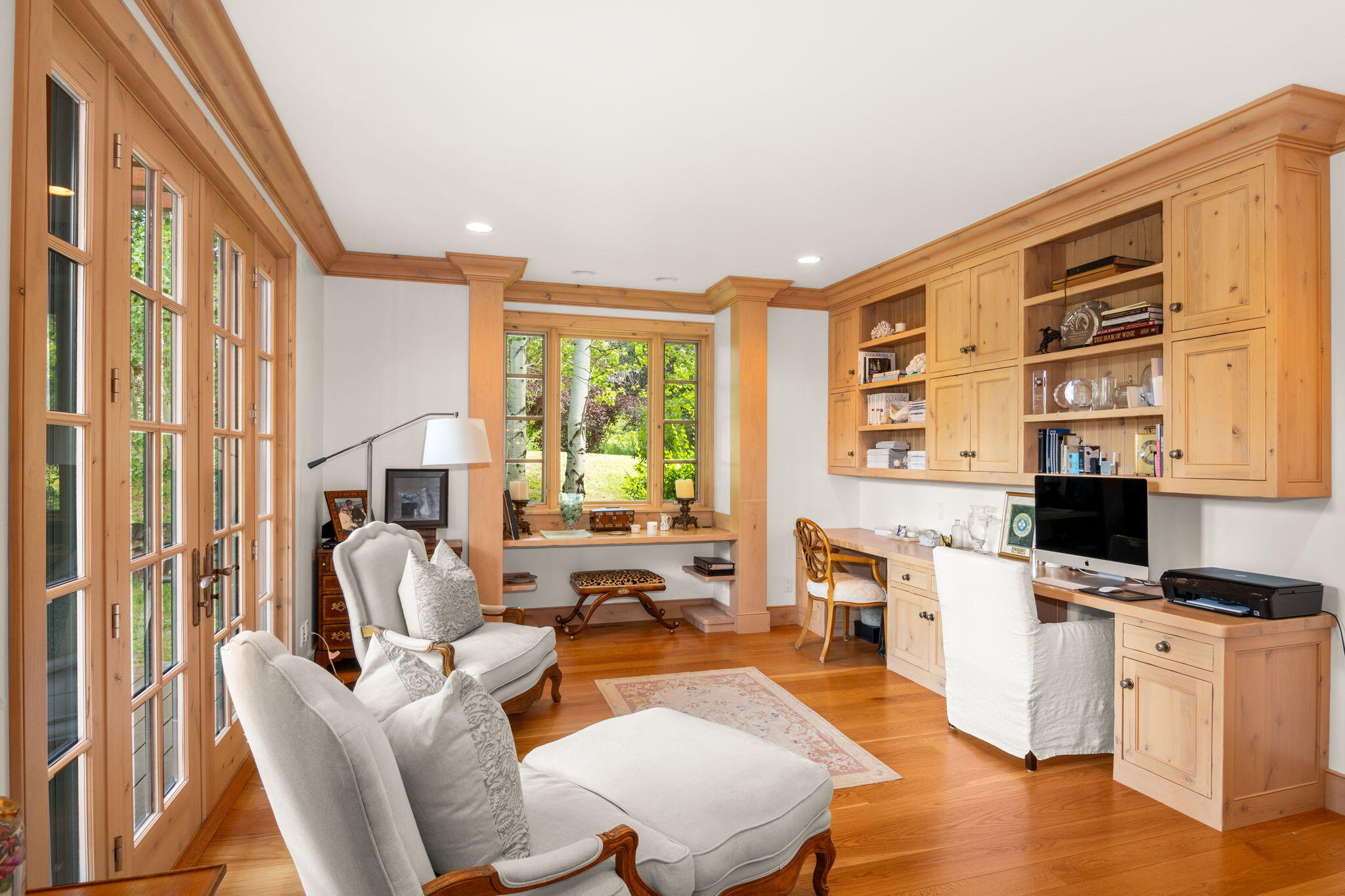 Home office with built-in wood cabinetry, a desk, white chairs, and tall garden-facing windows.