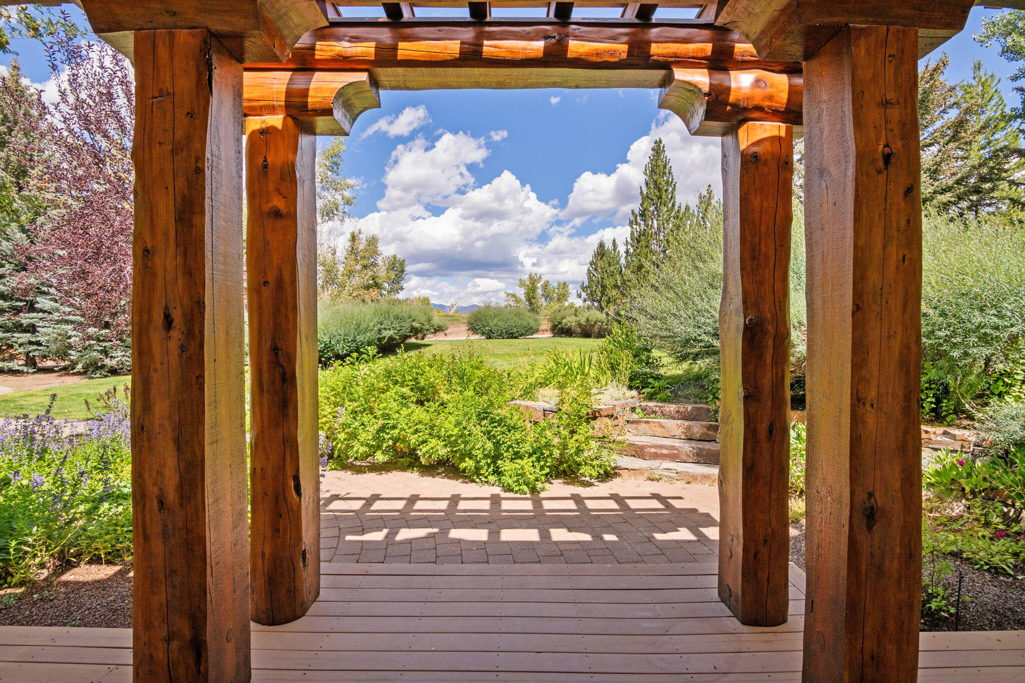 Timber pergola frames a garden patio with stone steps, flowering shrubs, and distant mountains.