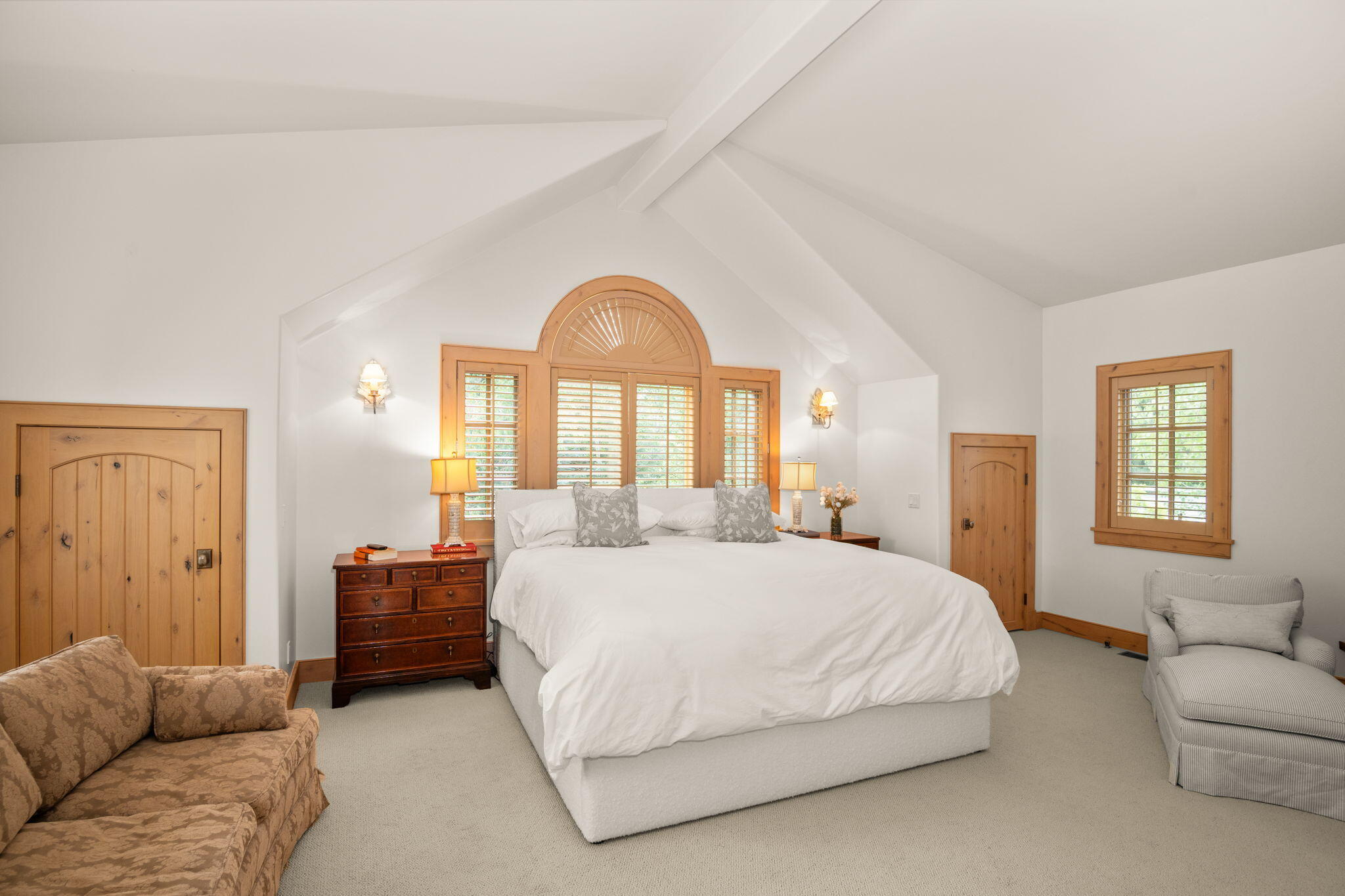 Furnished bedroom with a white bed, vaulted ceiling, and arched wood shutters.