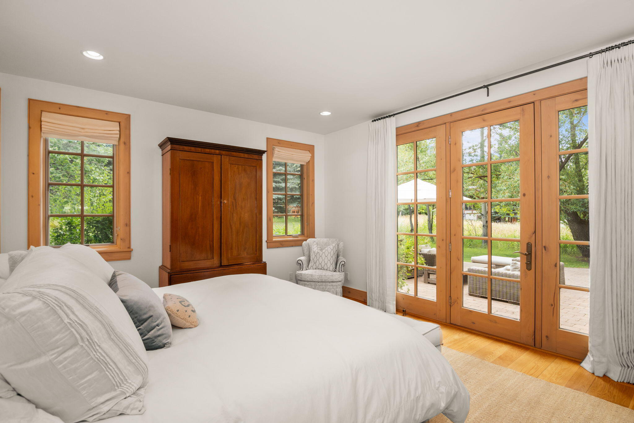 Bedroom with a white bed and wood-framed French doors opening onto a leafy patio.
