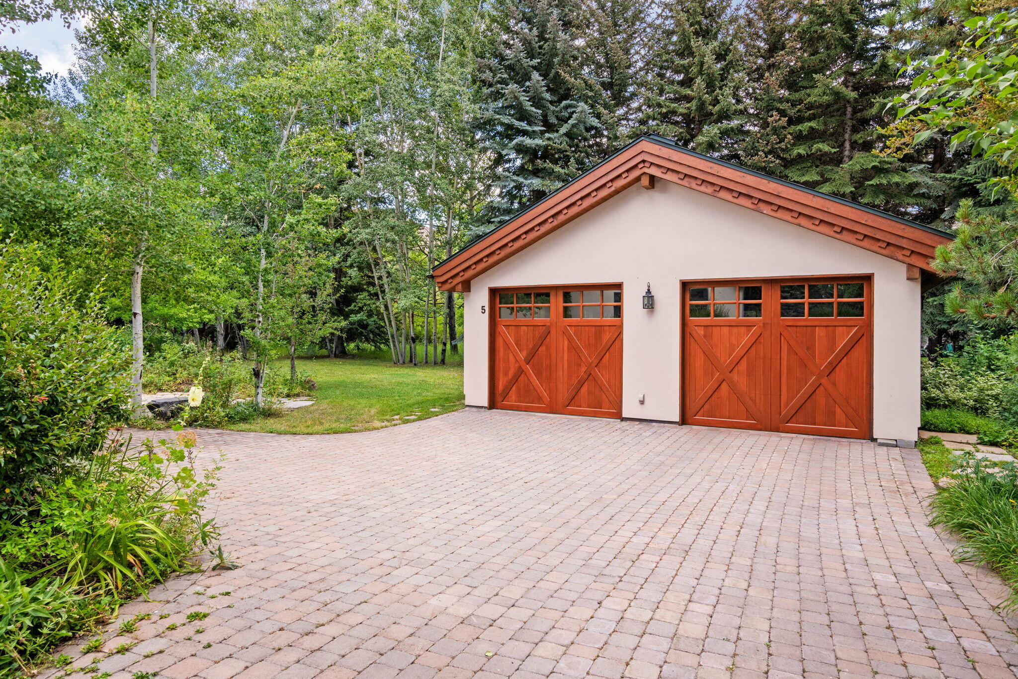 Detached garage with reddish-brown wooden doors and a broad paved driveway, surrounded by mature trees.