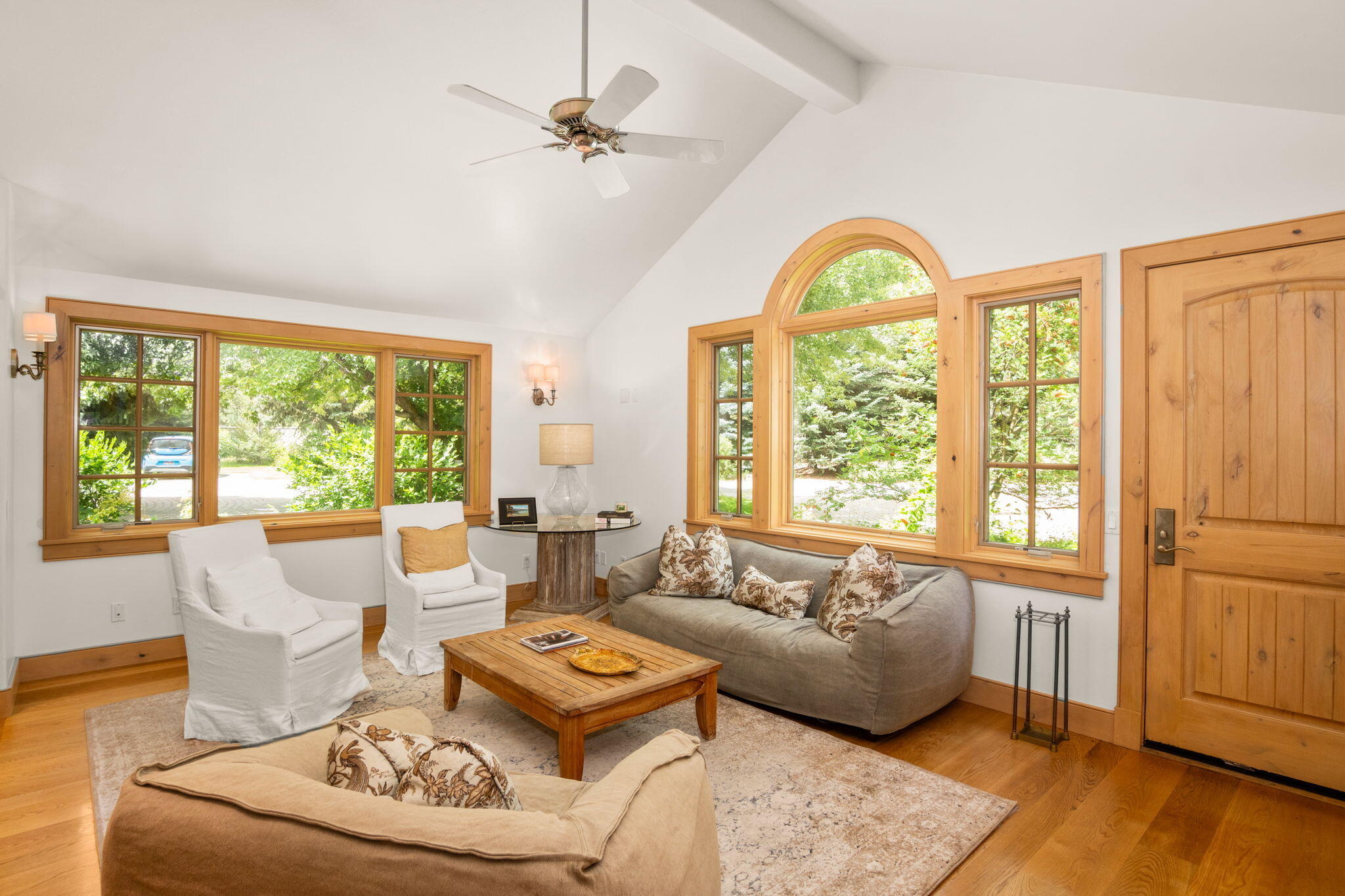 Living room with vaulted ceiling, wood-trimmed windows, gray sofa, and white armchairs.