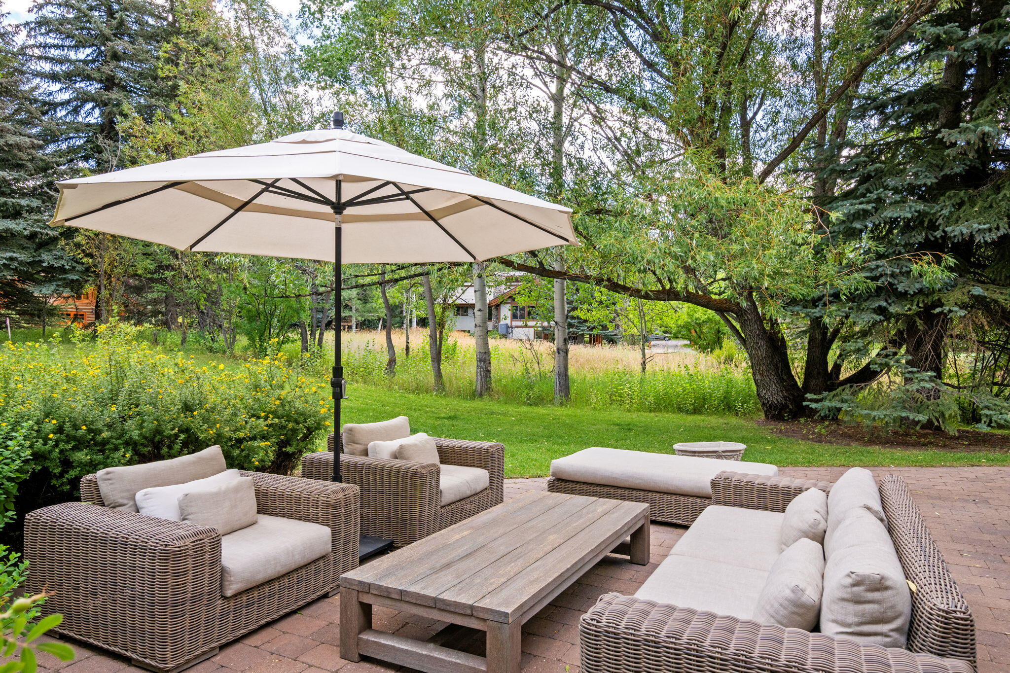 Outdoor patio with wicker seating and a large umbrella beneath leafy trees.