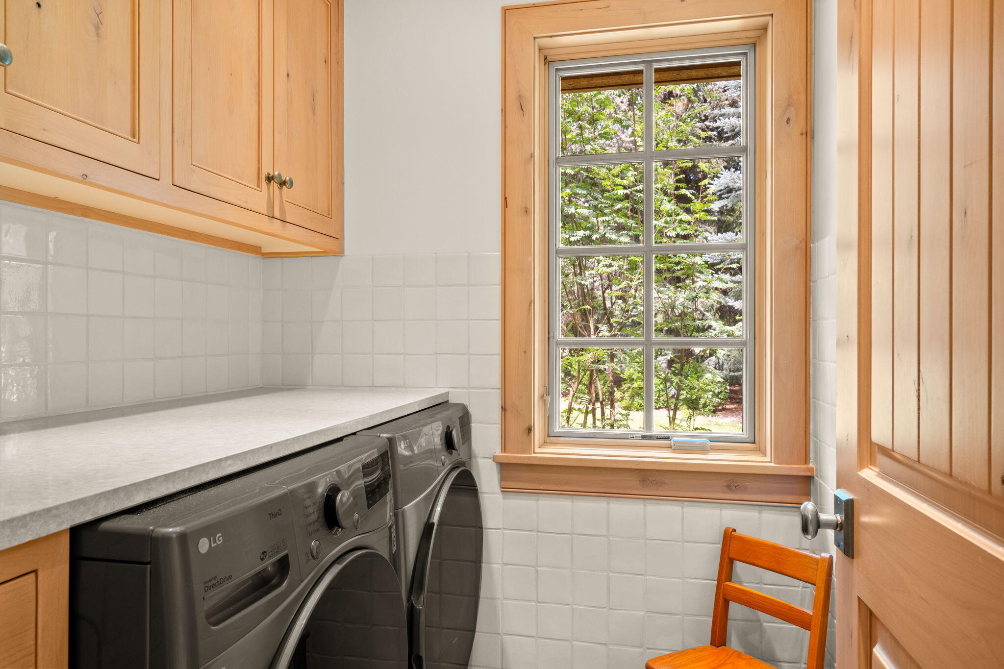 Laundry room with front-loading machines, light wood cabinets, and a window overlooking leafy trees.