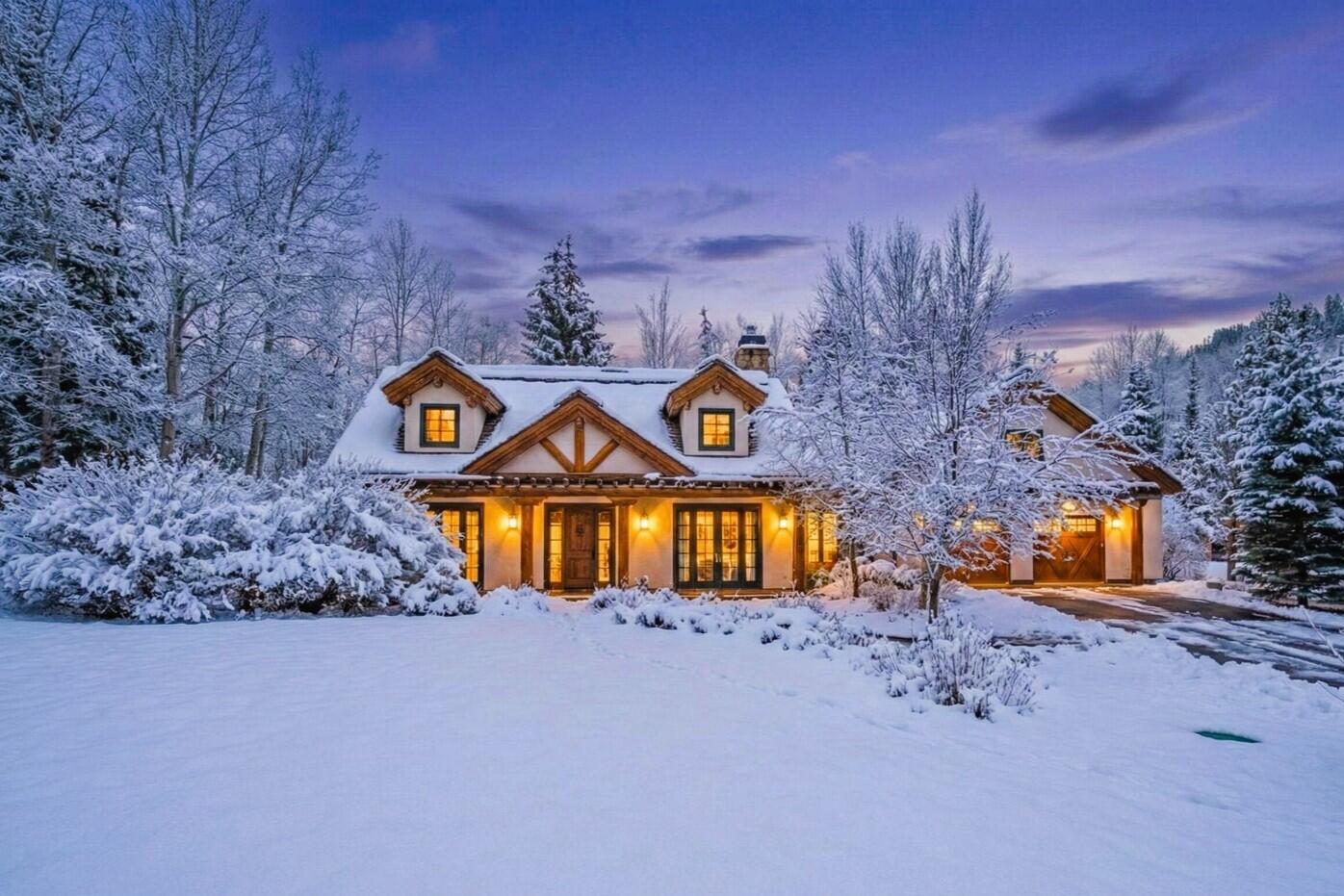 Snow-covered home with timber gables and warmly lit windows among frosted trees at dusk.