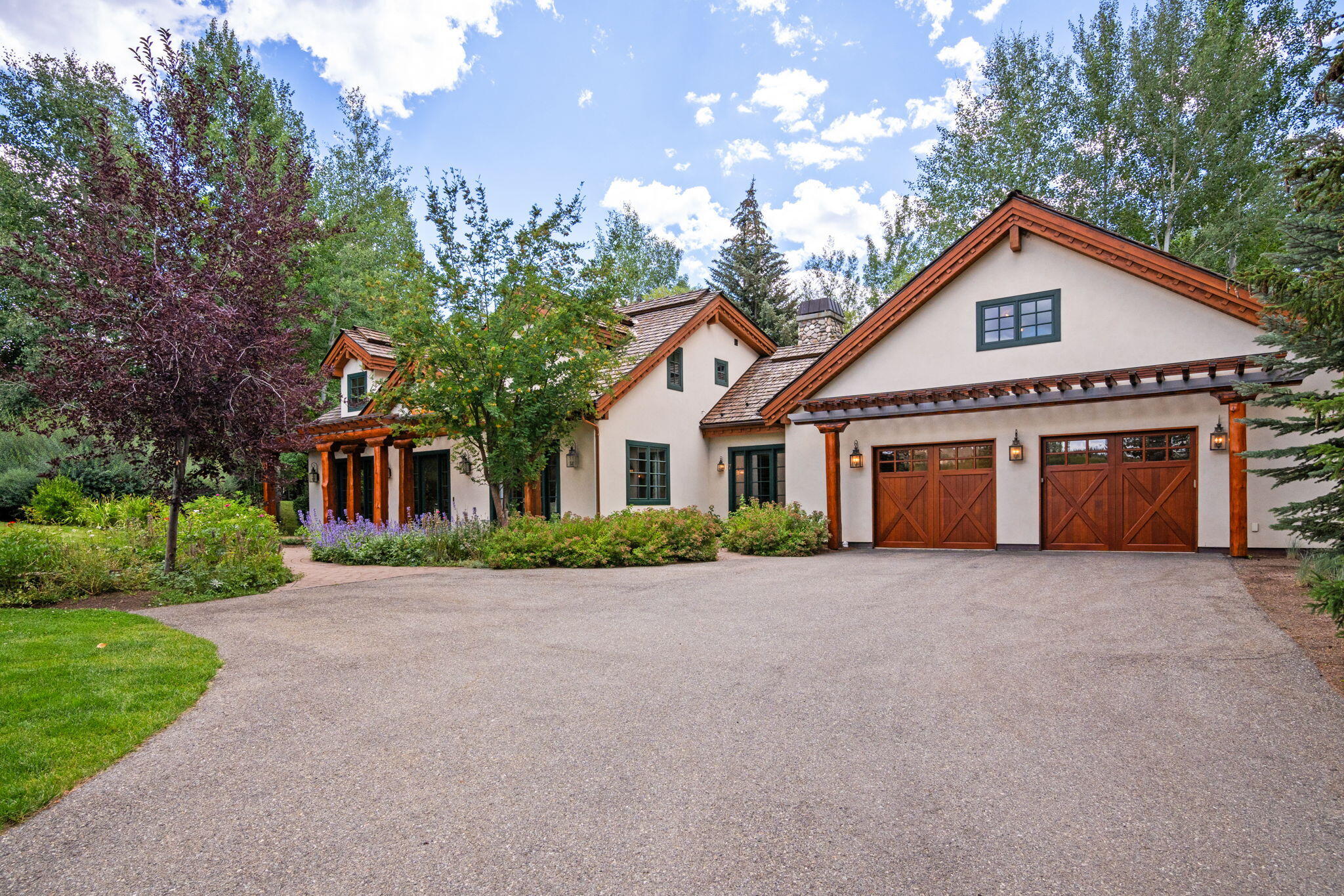 Stucco home with timber-trimmed gables and two wooden garage doors, framed by mature trees.