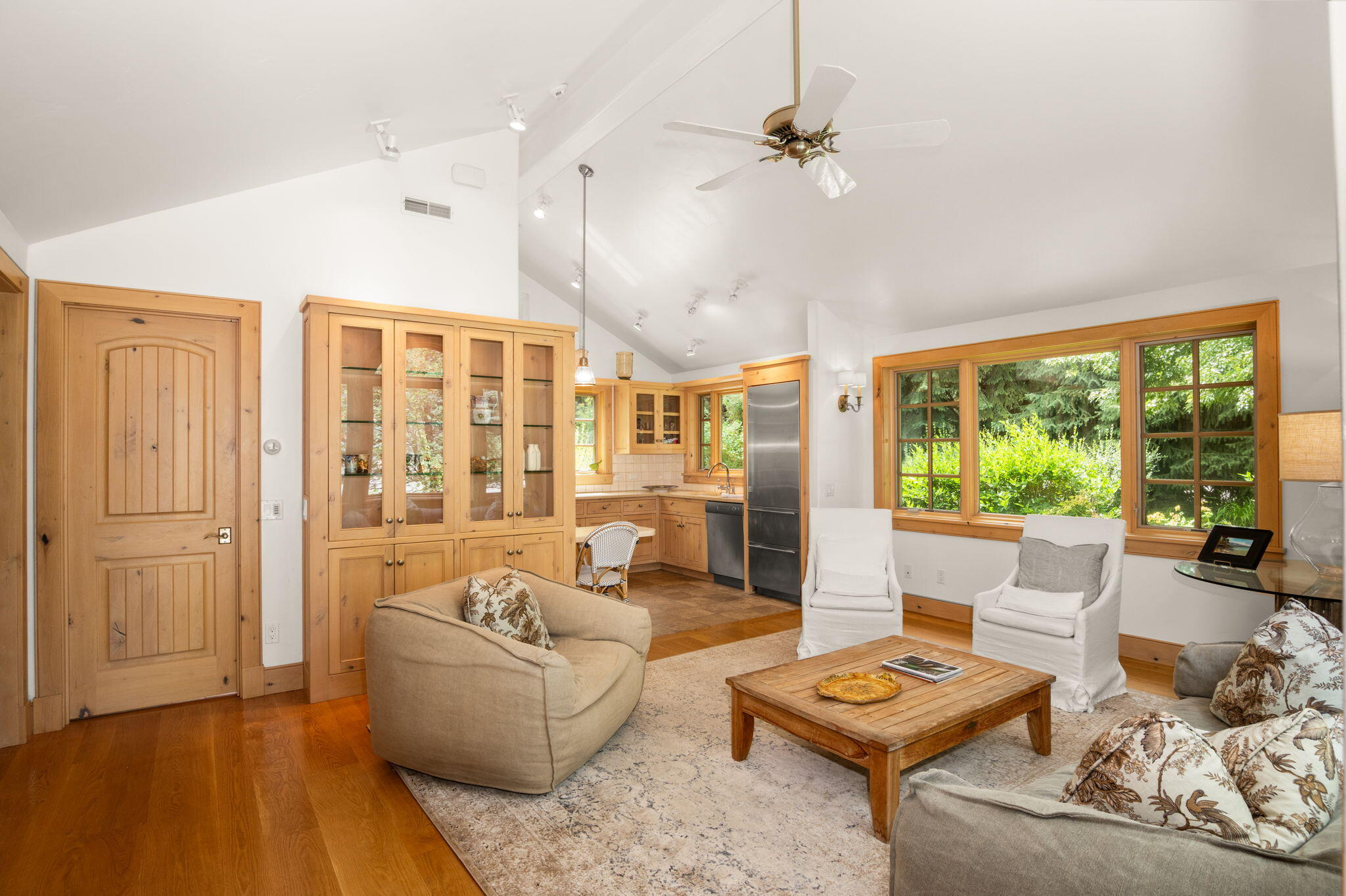 Open-plan living room and kitchen with vaulted ceiling, wood cabinetry, and large windows overlooking greenery.