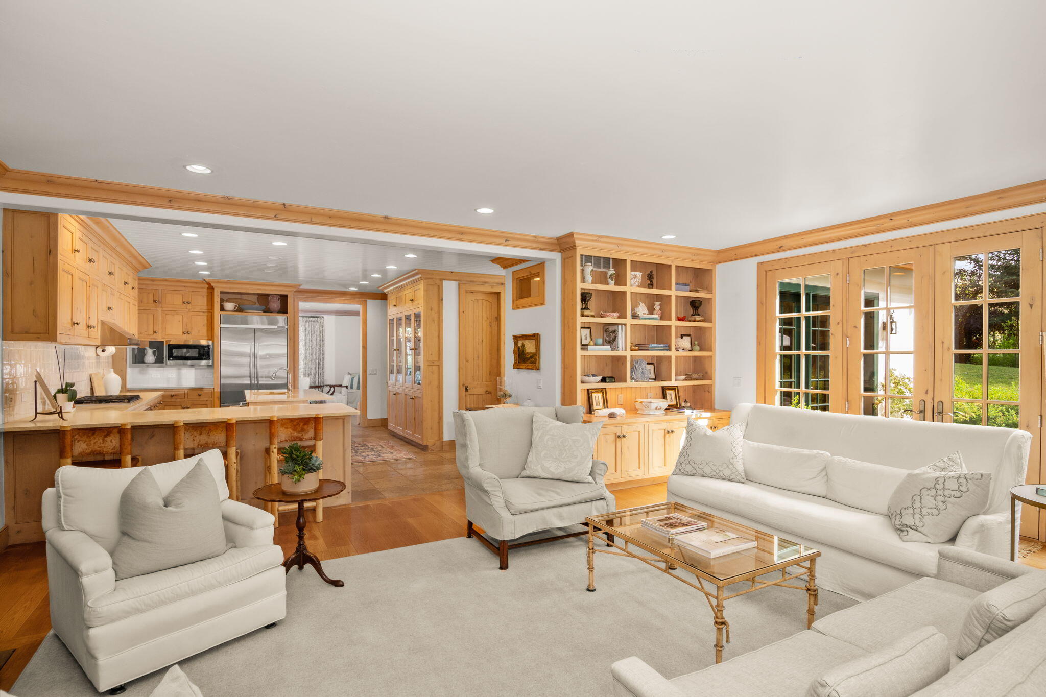Living room with white seating, built-in wood shelves, an open kitchen, and French doors to the lawn.
