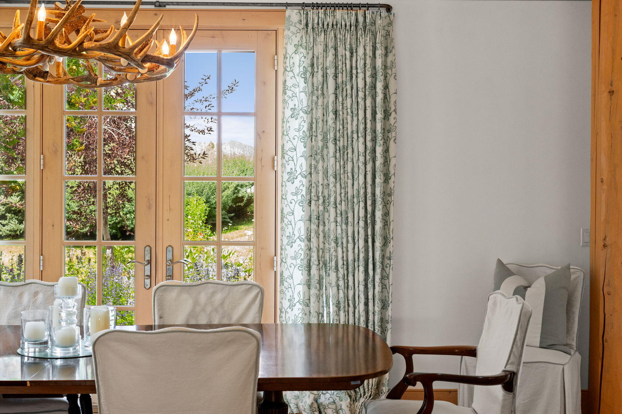 Dining room with an antler chandelier and French doors overlooking a garden and distant mountain.