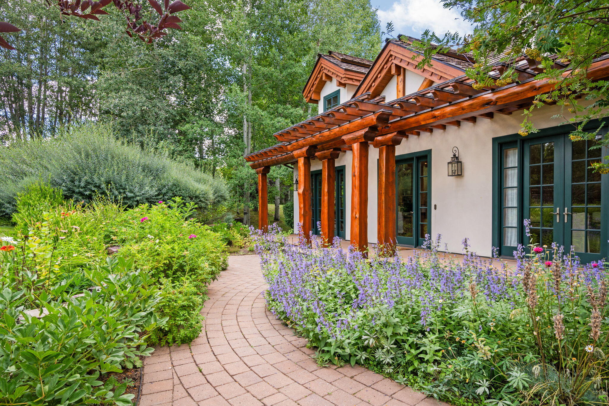 House exterior with timber pergola and French doors beside a curved path through flowering gardens.
