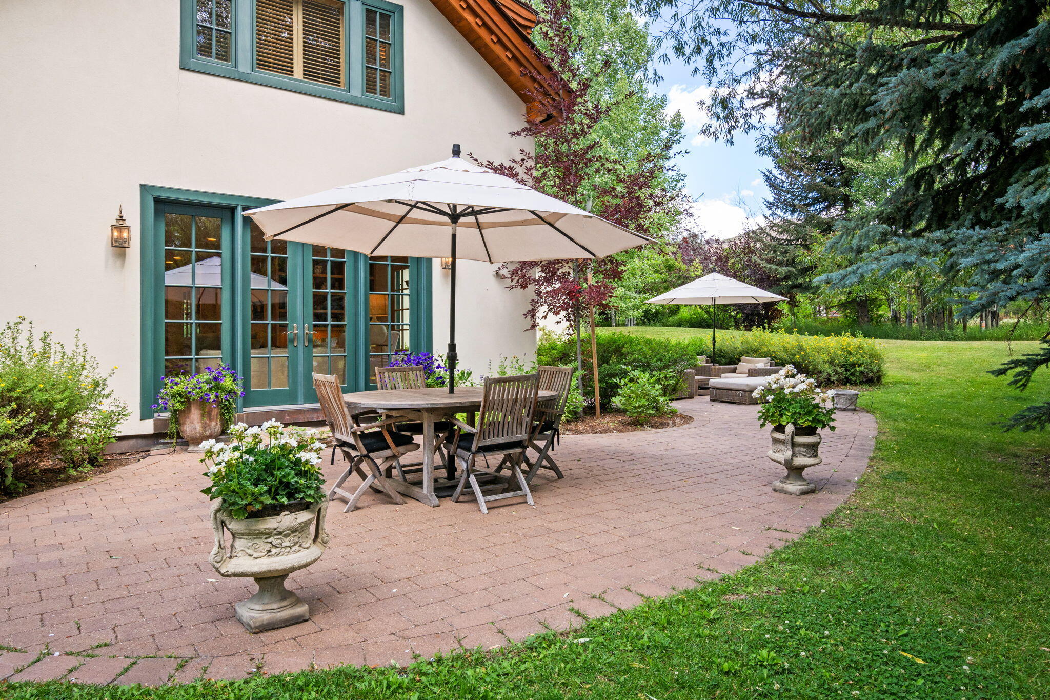Brick patio with teal-framed doors, umbrella-covered dining table, flower planters, and garden lounge seating.