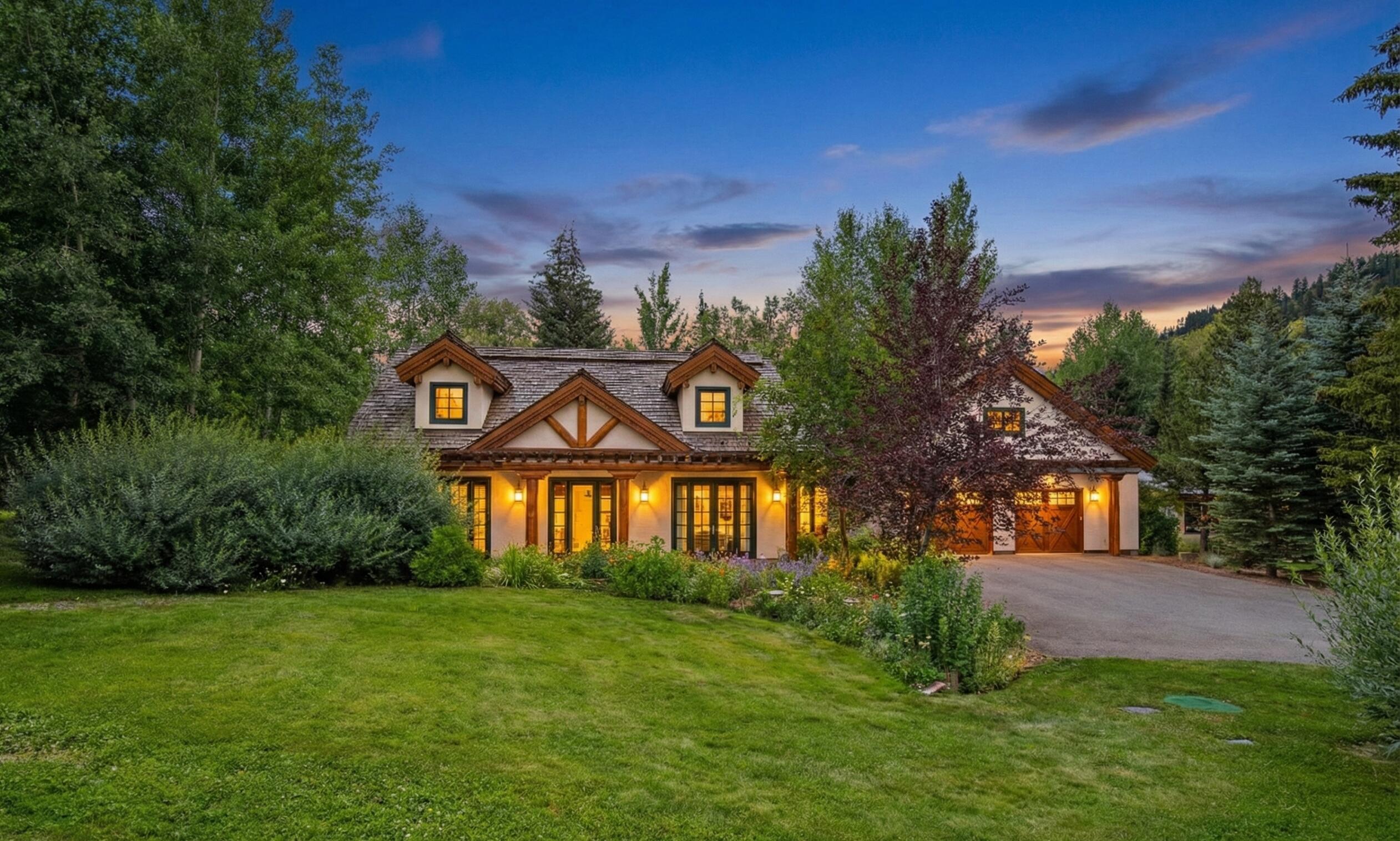 Two-story home with timber trim and lit windows, surrounded by mature trees and garden beds at dusk.