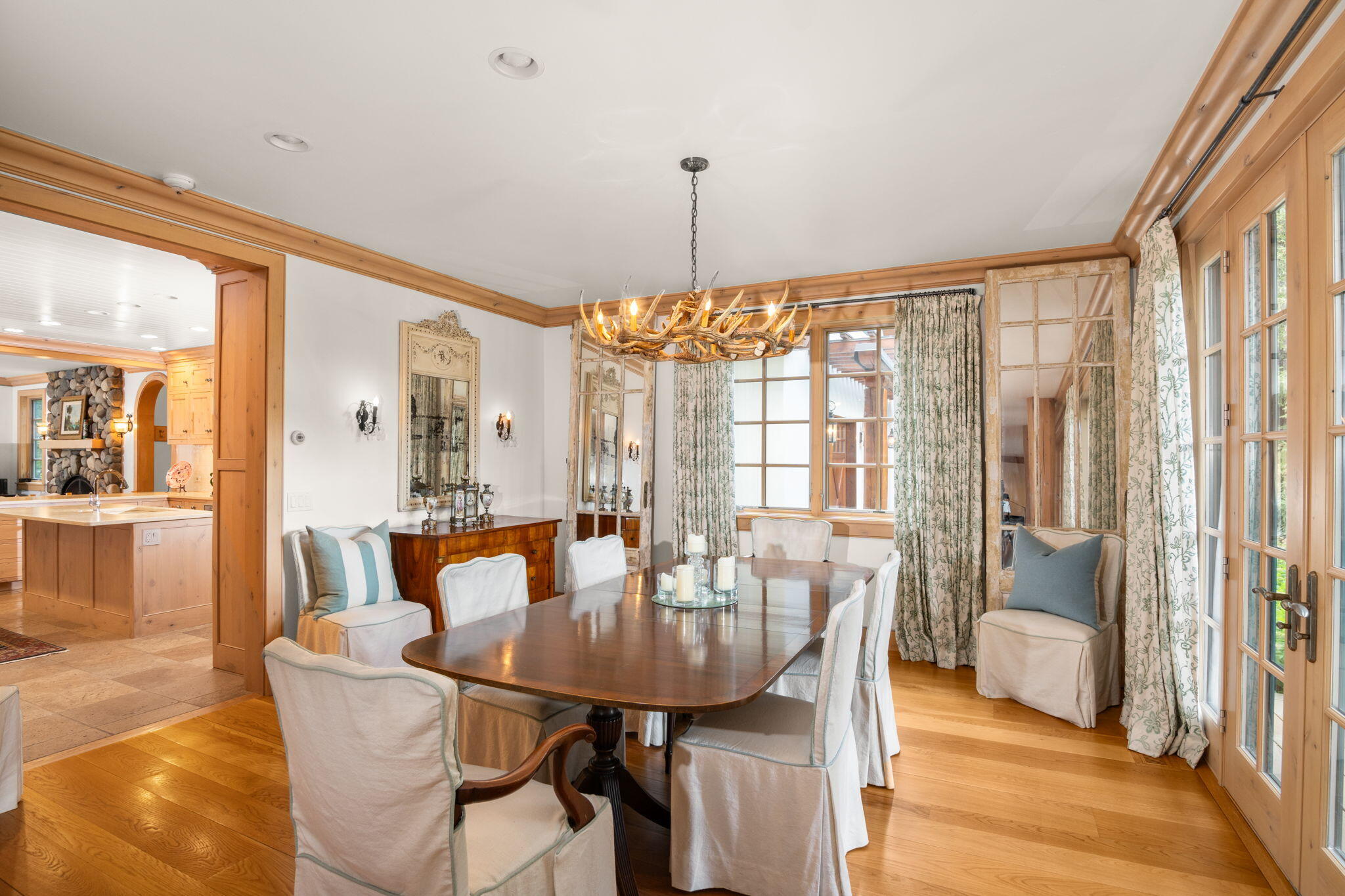 Dining room with an antler chandelier, wood table, tall windows, and patterned curtains.