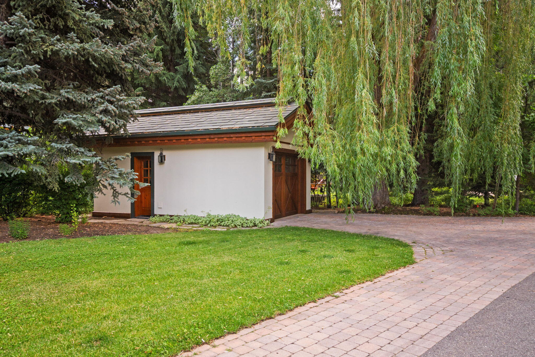 Detached garage with white stucco walls and wood doors beside a curved brick driveway.