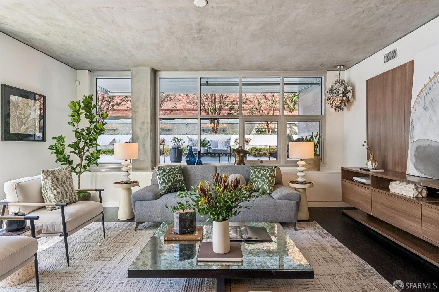 Staged living room with a gray sofa, glass coffee table, and large courtyard-facing windows.