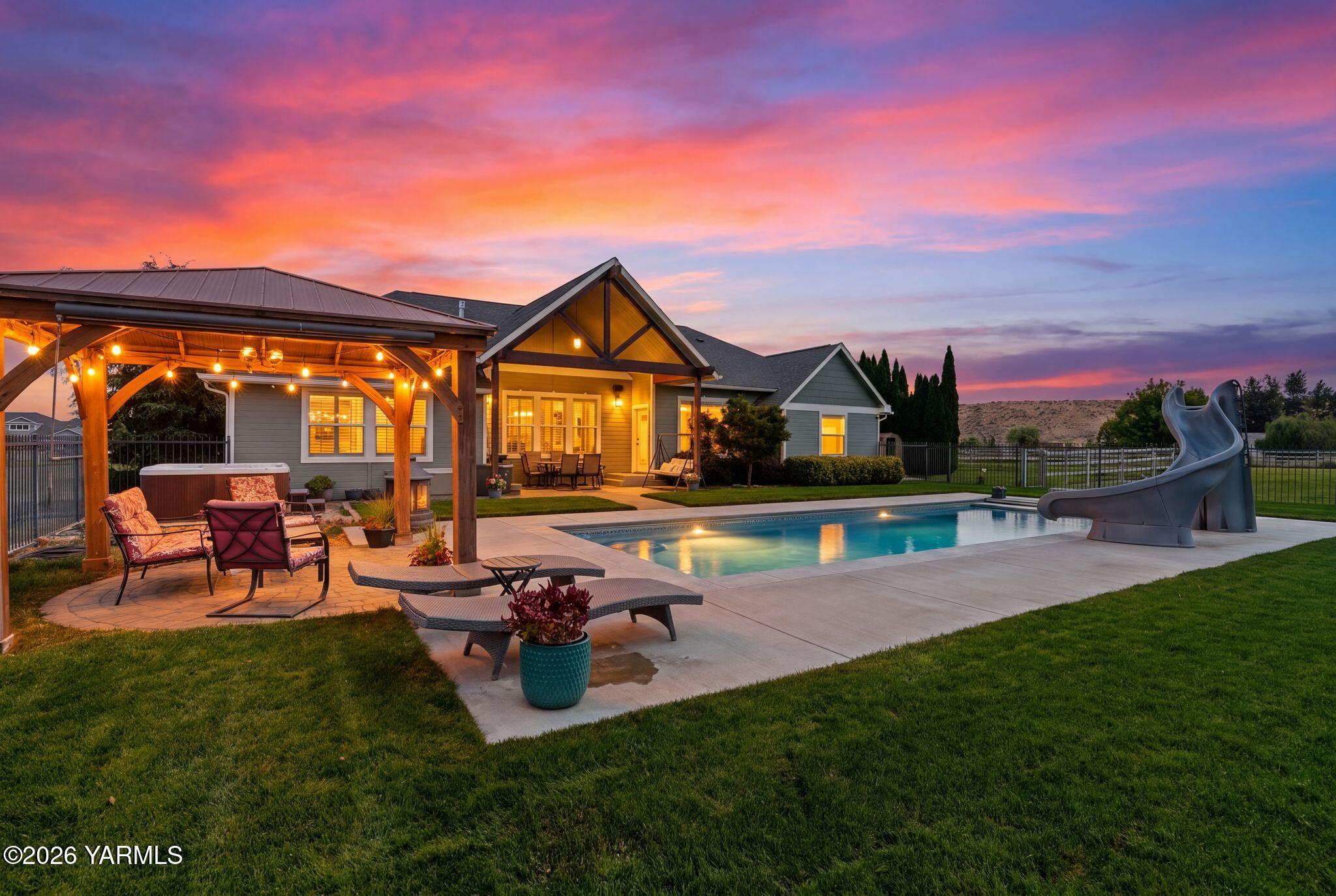 Home backyard at dusk with a lit covered patio, swimming pool, and large slide.