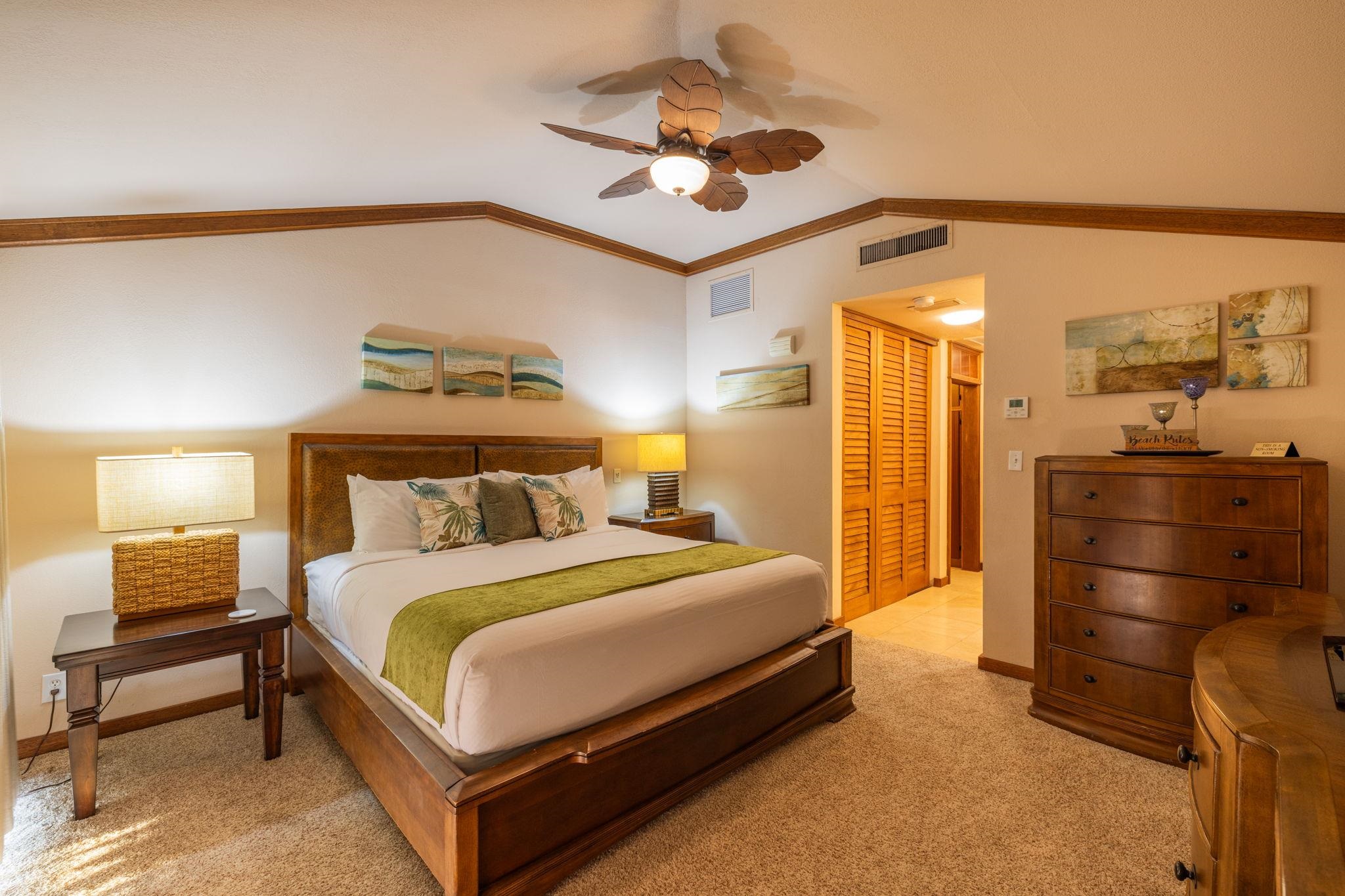 Furnished bedroom with a wooden bed, green runner, tropical pillows, and a leaf-shaped ceiling fan.