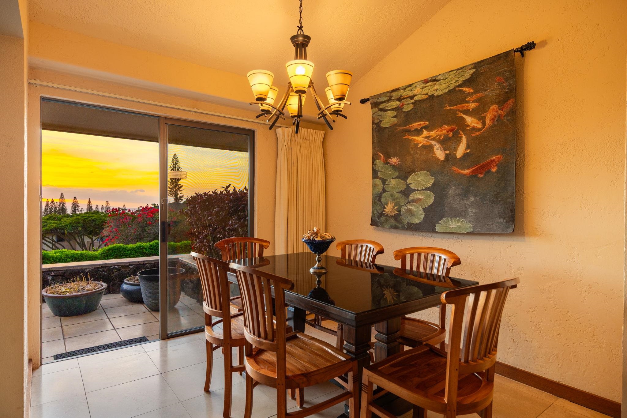 Dining room with a glass-top table and wooden chairs opening onto a garden patio at sunset.