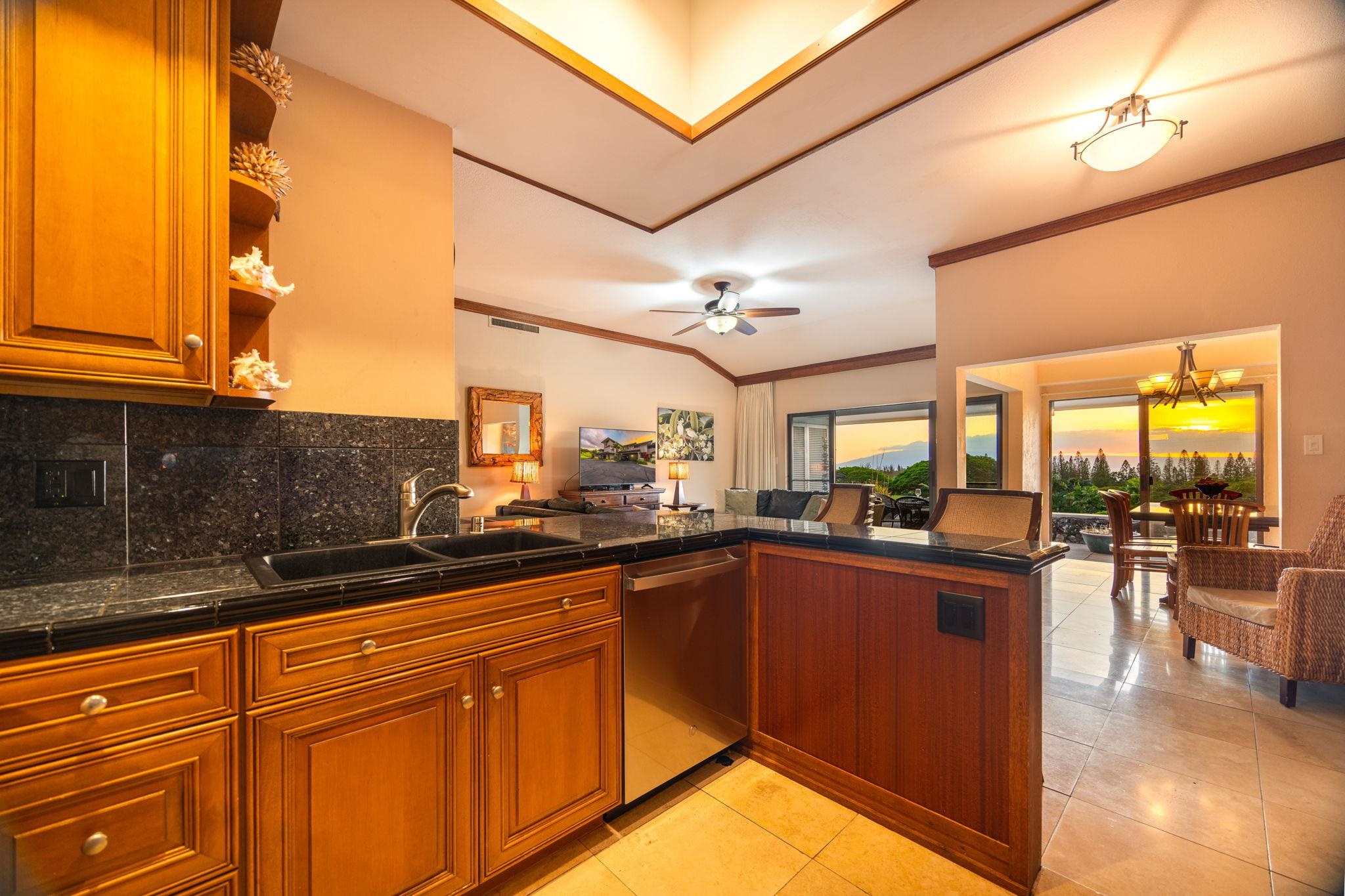 Open-plan kitchen with rich wood cabinets and stone island, looking toward a dining area and ocean sunset.