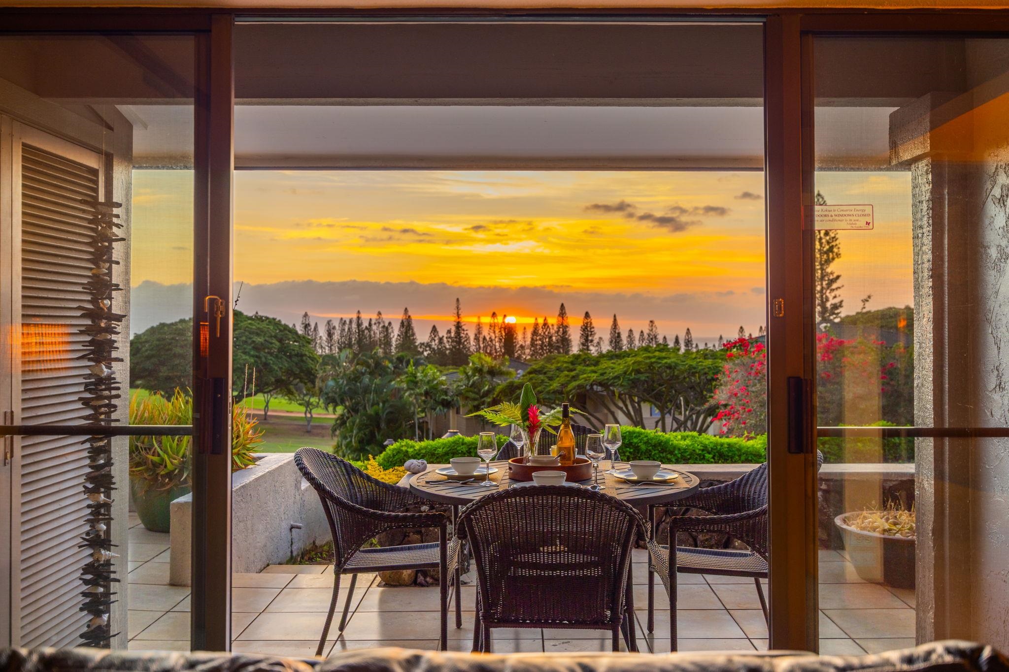 Patio dining table and wicker chairs overlook tropical gardens and a sunset over the water.