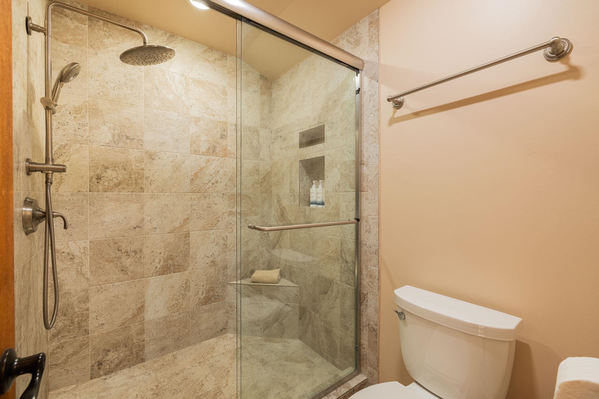 Stone-tiled bathroom shower with a glass enclosure, rainfall showerhead, and handheld sprayer.