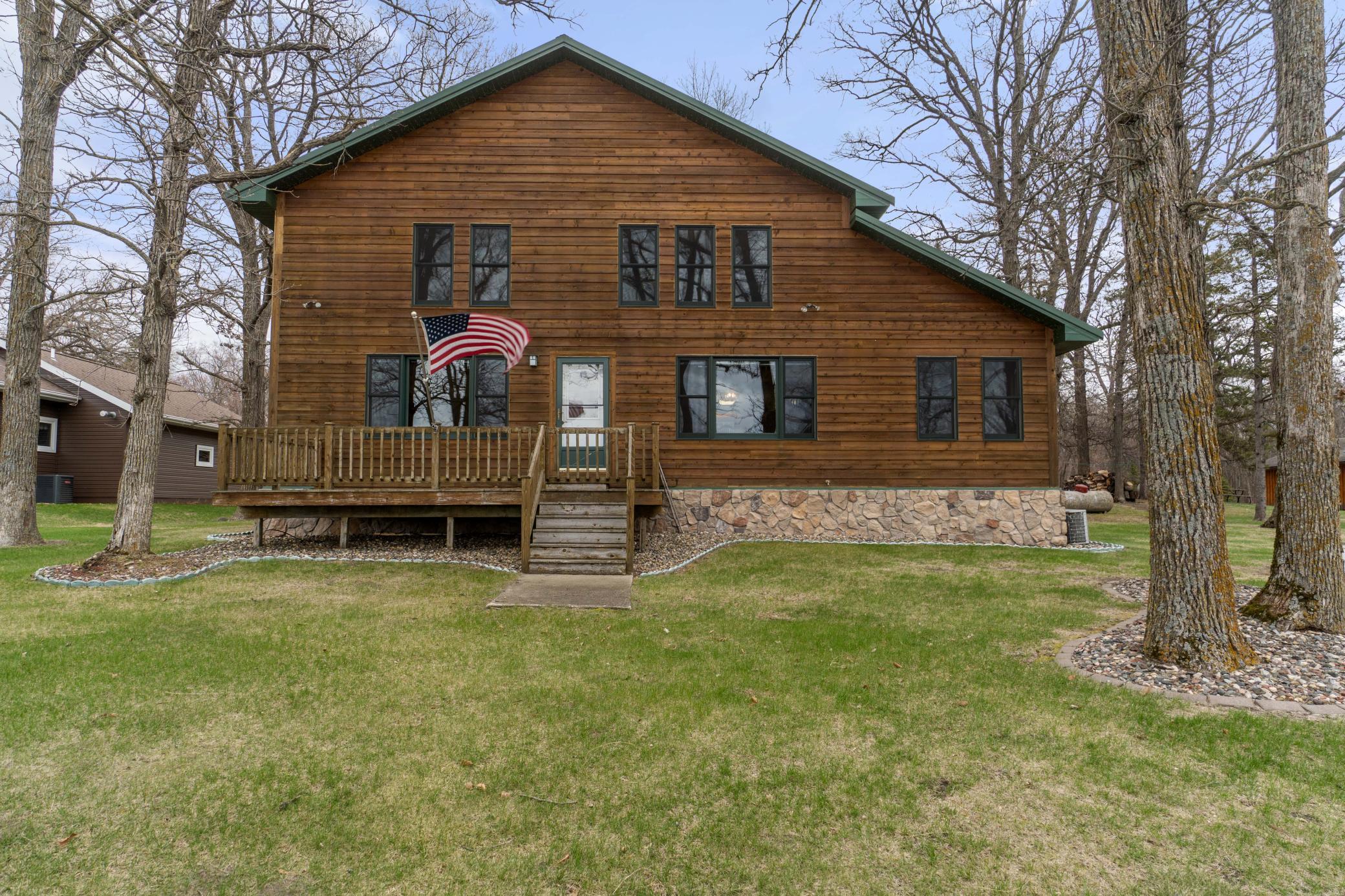 Wood-sided two-story home with a raised deck, stone foundation, and American flag.