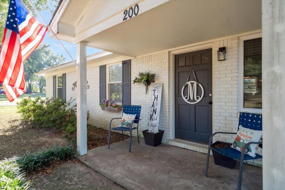 White-brick home’s covered front porch with a dark entry door, blue chairs, and an American flag.