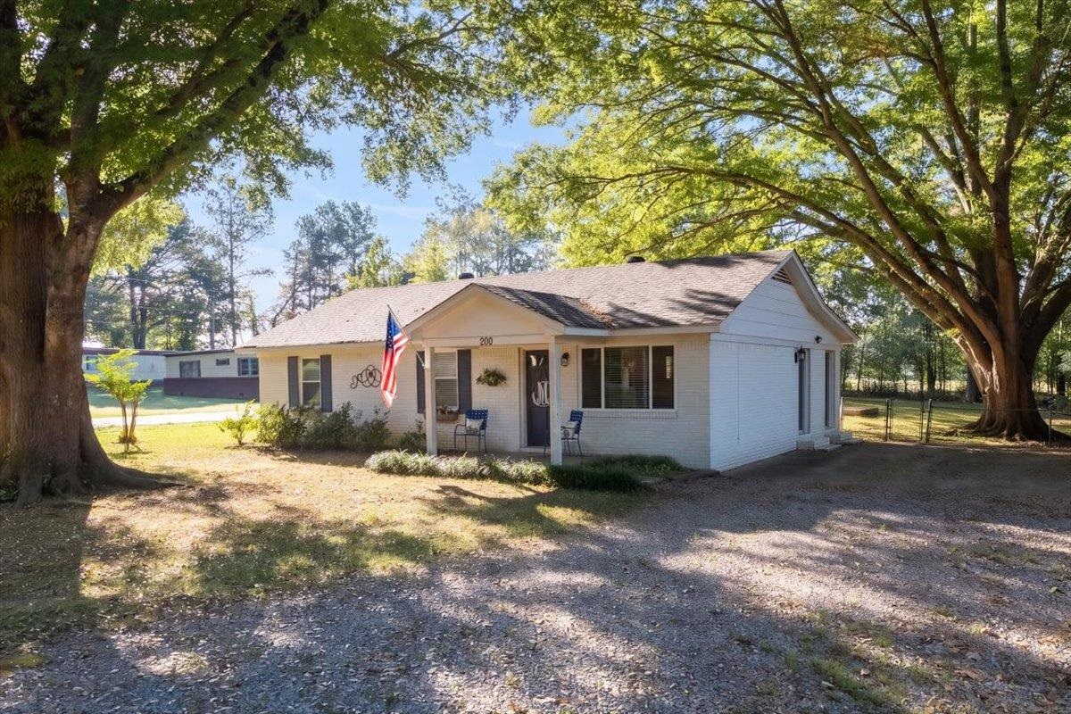 Single-story white house beneath large shade trees, with a gravel driveway and American flag.