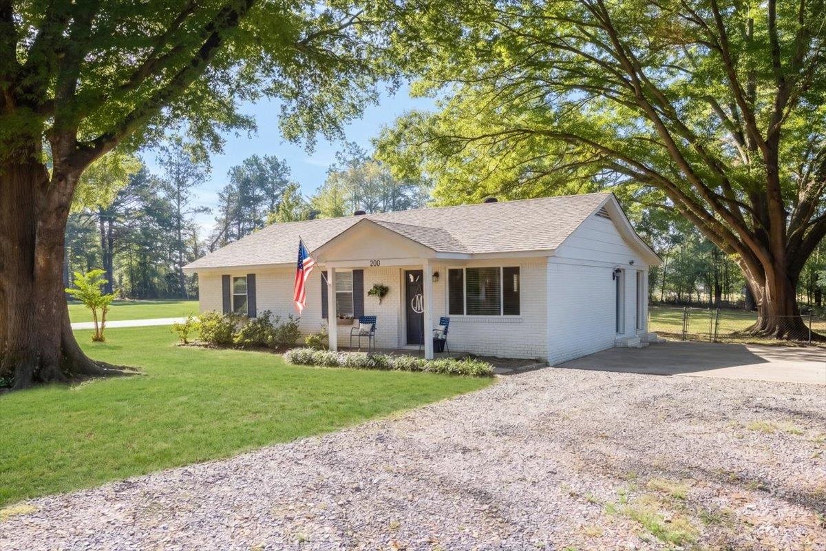 Single-story white brick home beneath large shade trees, with a gravel drive and American flag.