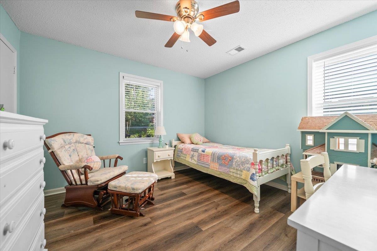 Bedroom with a white bed, floral rocking chair, and oversized dollhouse beside bright windows.