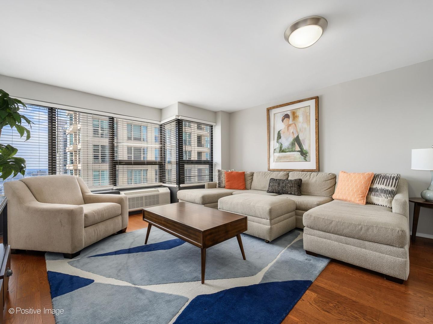 Furnished condo living room with a beige sectional, blue geometric rug, and corner windows.