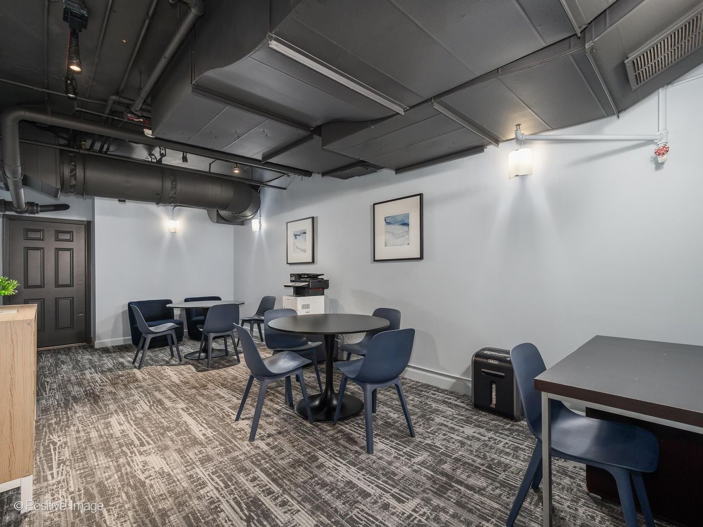 Communal meeting room with round tables, blue chairs, and exposed ceiling ducts.