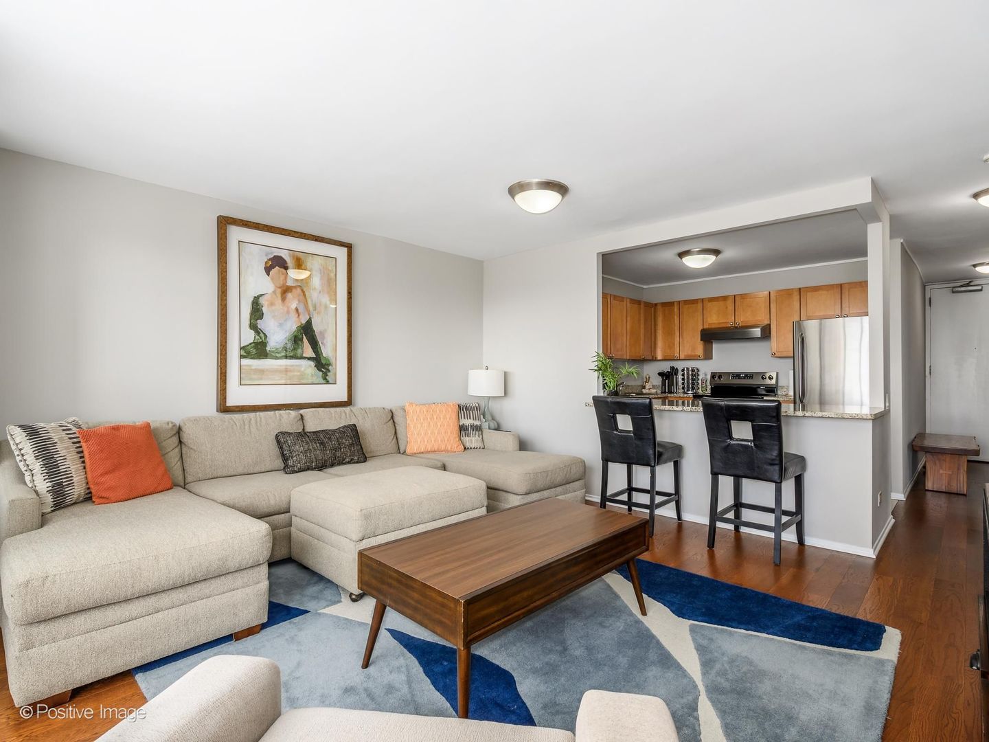 Furnished condo living room with a beige sectional and an open kitchen with wood cabinets.