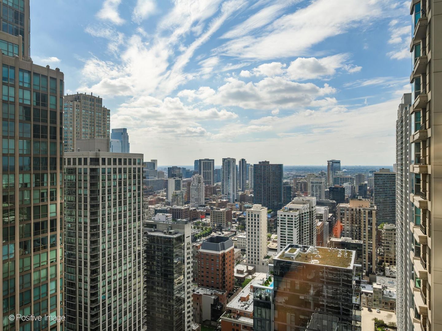 Downtown Chicago skyline viewed from above, framed by nearby towers beneath a bright, cloud-streaked sky.