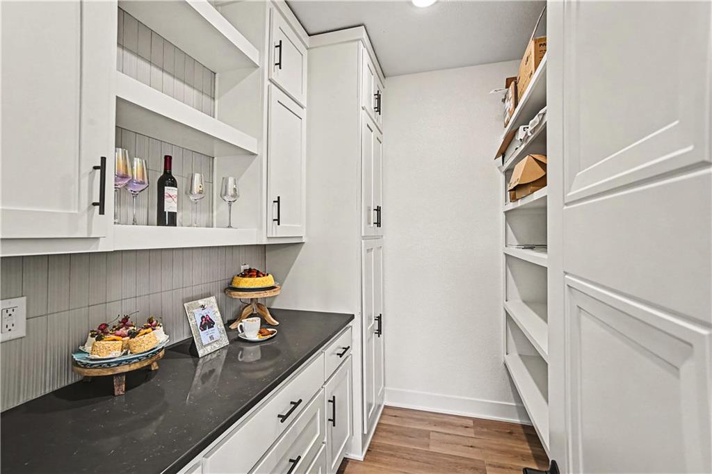 Walk-in pantry with white cabinets, dark countertop, gray tile backsplash, and wood-look flooring.