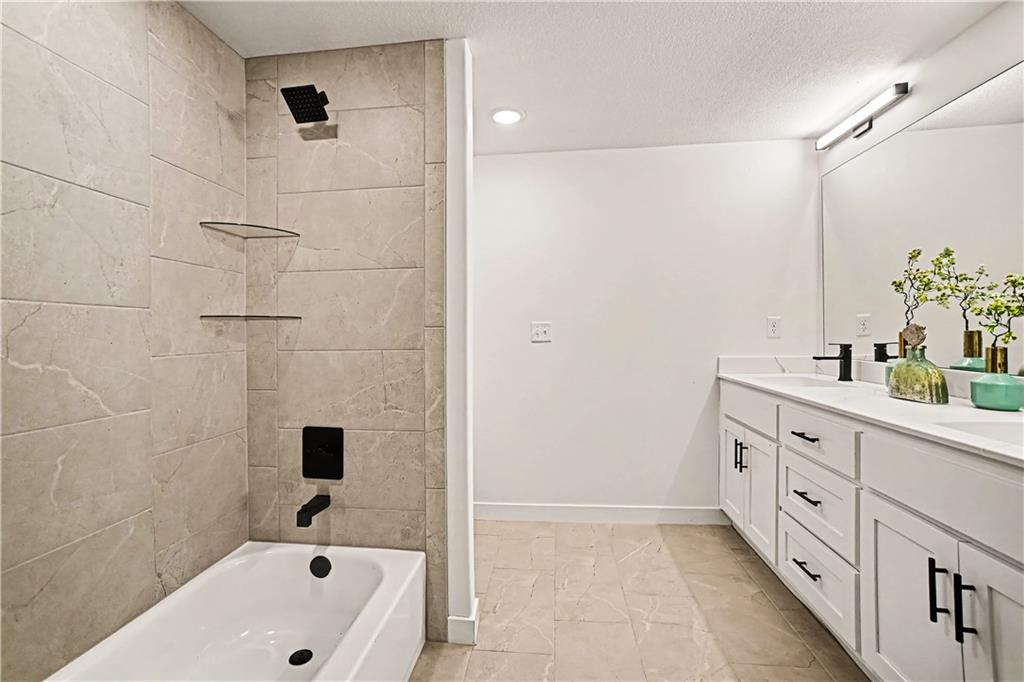 Bathroom with a beige stone-tiled shower, white tub, and long double vanity with black fixtures.