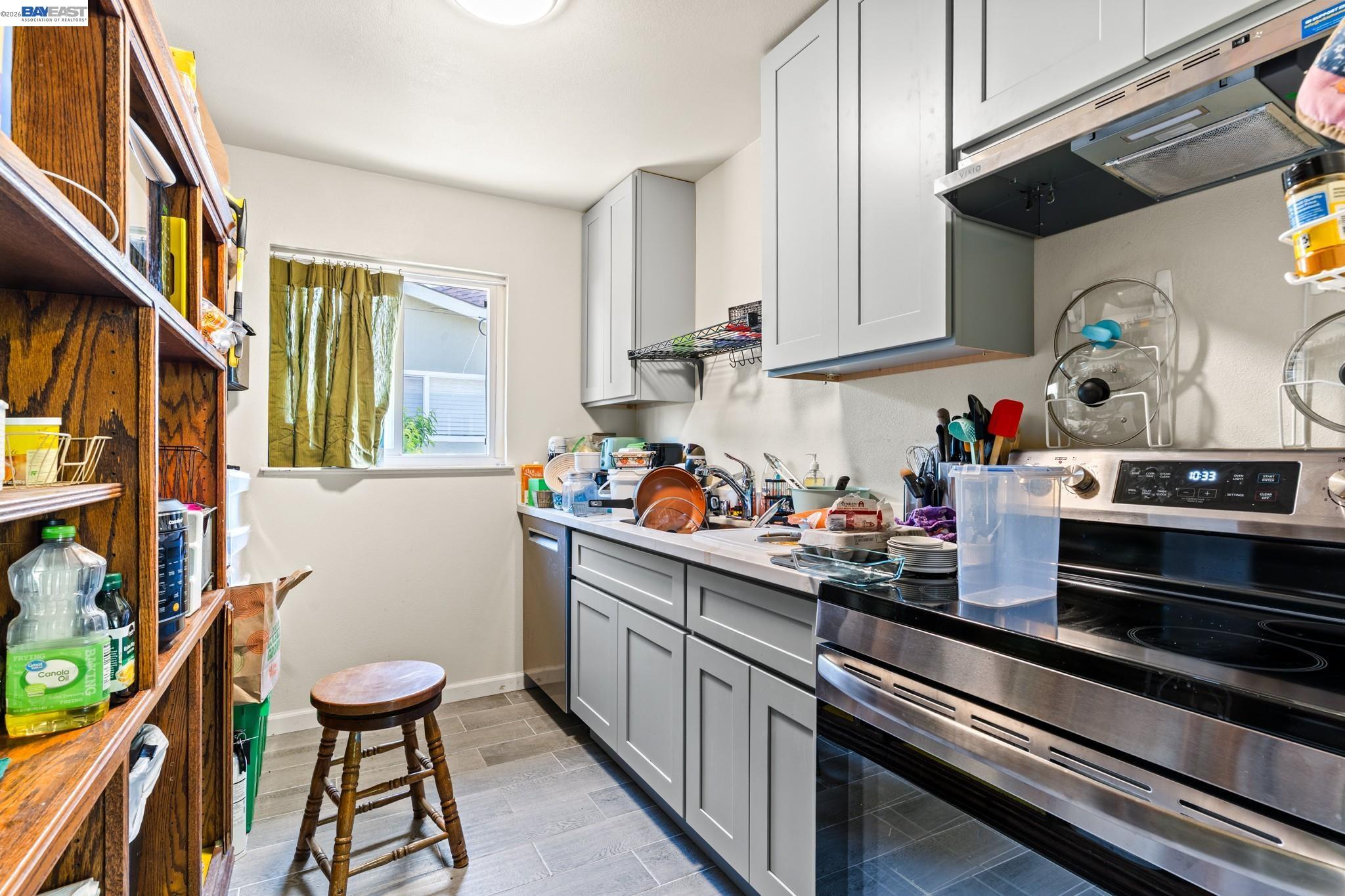 Kitchen with gray cabinets, stainless steel range, tiled floor, and crowded counters beside a curtained window.
