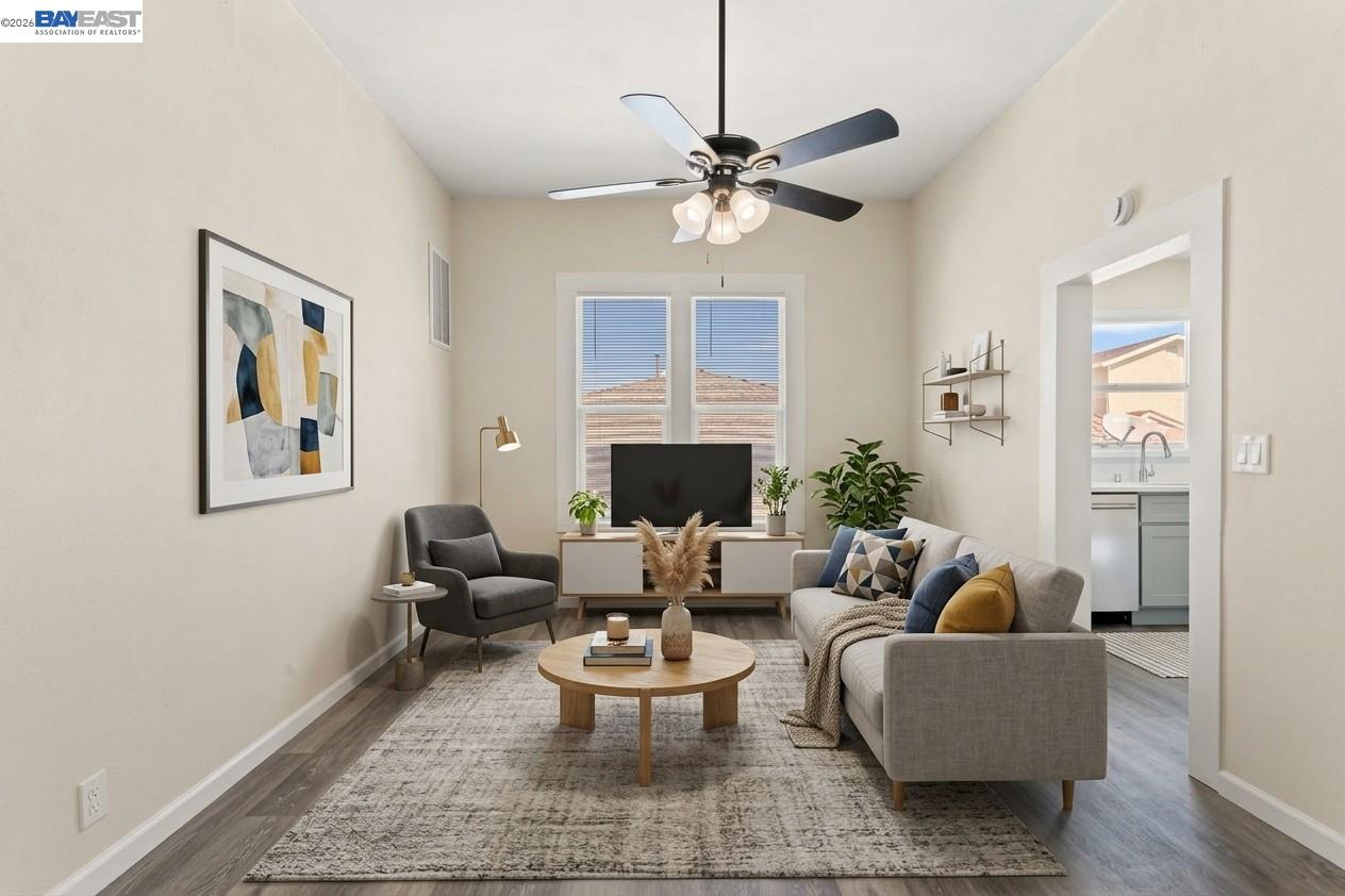 Furnished living room with a gray sofa and armchair, ceiling fan, and glimpse of the adjacent kitchen.
