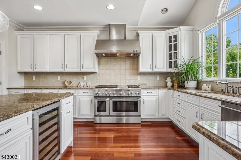 Kitchen with white cabinetry, granite counters, stainless double ovens, hardwood floors, and large windows.