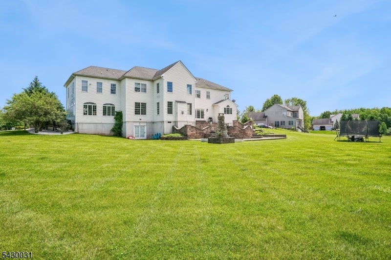 Large white two-story house beyond a broad lawn, with a stone patio and trampoline.